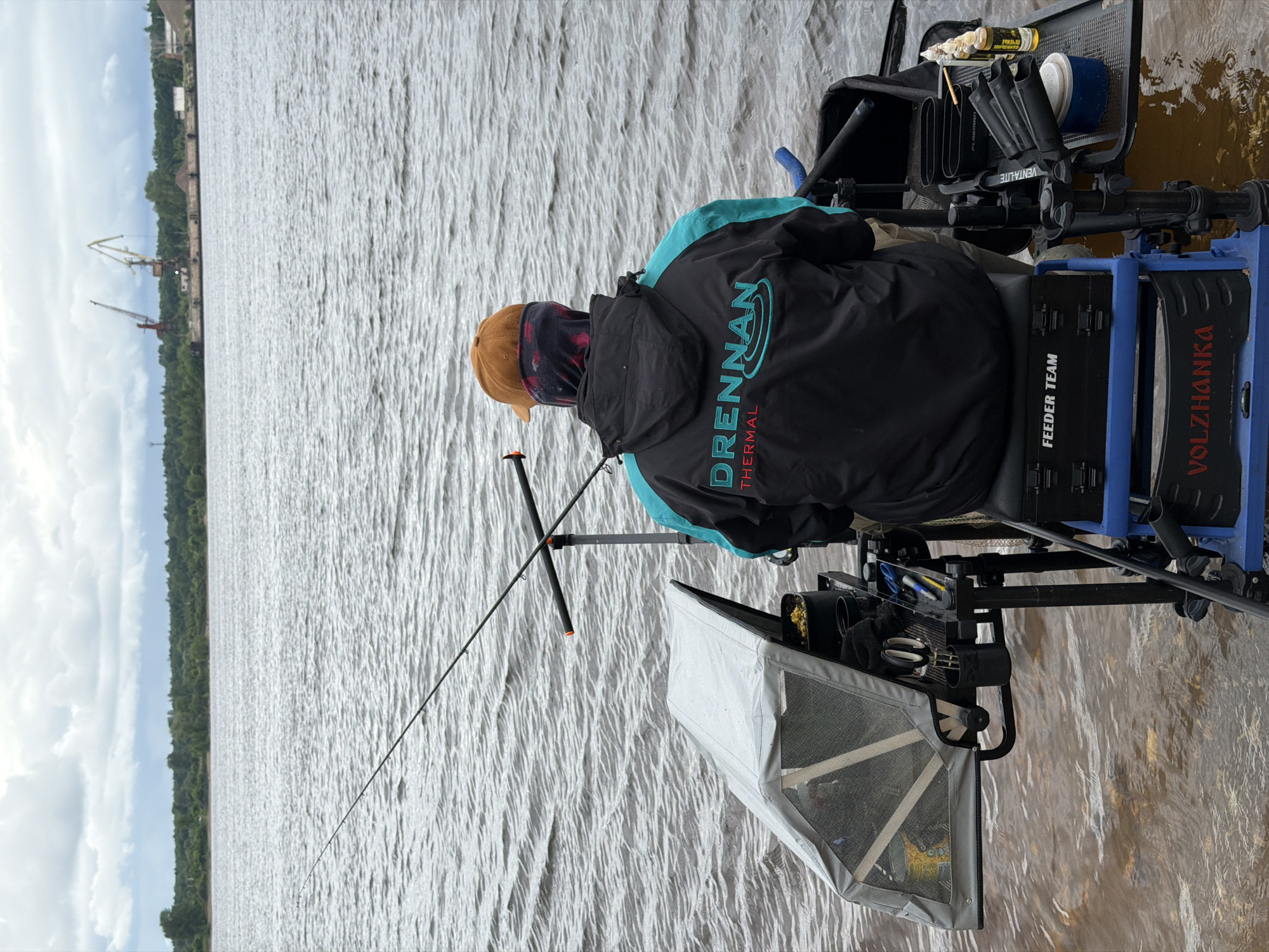 An angler is depicted from behind, seated in a specialized fishing setup in the shallow, brownish water of a wide river under an overcast sky in Chaikovsky, Russia. The individual wears a black jacket with teal sleeves, bearing "DRENNAN THERMAL" branding, a brown baseball cap, and a multicolored neck gaiter. They are seated on a blue and black fishing seatbox, with a black "FEEDER TEAM" storage box integrated into the seat. A single fishing rod extends diagonally from the right into the water, resting on an adjustable rod support. To the angler's left, a grey canopy with mesh panels covers a container holding yellow bait (likely corn), positioned on an attached side tray that also holds scissors, pens, and other small items. To the angler's right, another side tray displays several bottles of liquid attractants, some with "FLAGMAN" branding, a blue container, and a multi-slot rod holder. The river's surface is textured with ripples. The distant bank features a dense treeline. To the right on the far bank, two large industrial cranes, one red and one yellow, are visible above a dock or industrial embankment with associated structures.