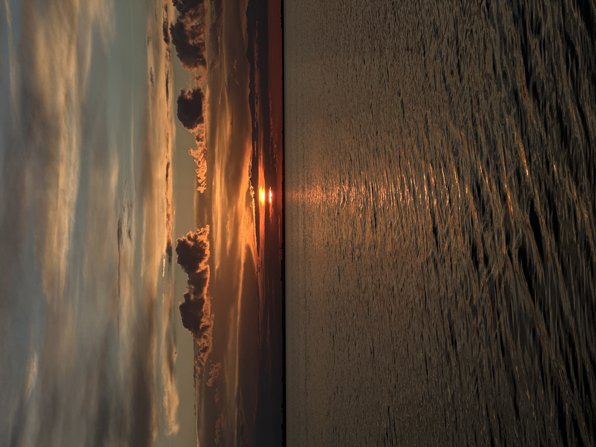 This is a photograph of a sunset over a body of water. The sky is filled with dramatic clouds in shades of orange, red, and gray, with the sun peeking through on the horizon. The water reflects the colors of the sunset, creating a shimmering path of light. The horizon line is low, with a dark silhouette of land visible in the distance. The overall mood of the image is serene and atmospheric. It is not possible to determine the specific location within the city of Kokshetau, Kazakhstan, or the presence of people or specific objects from this image alone.