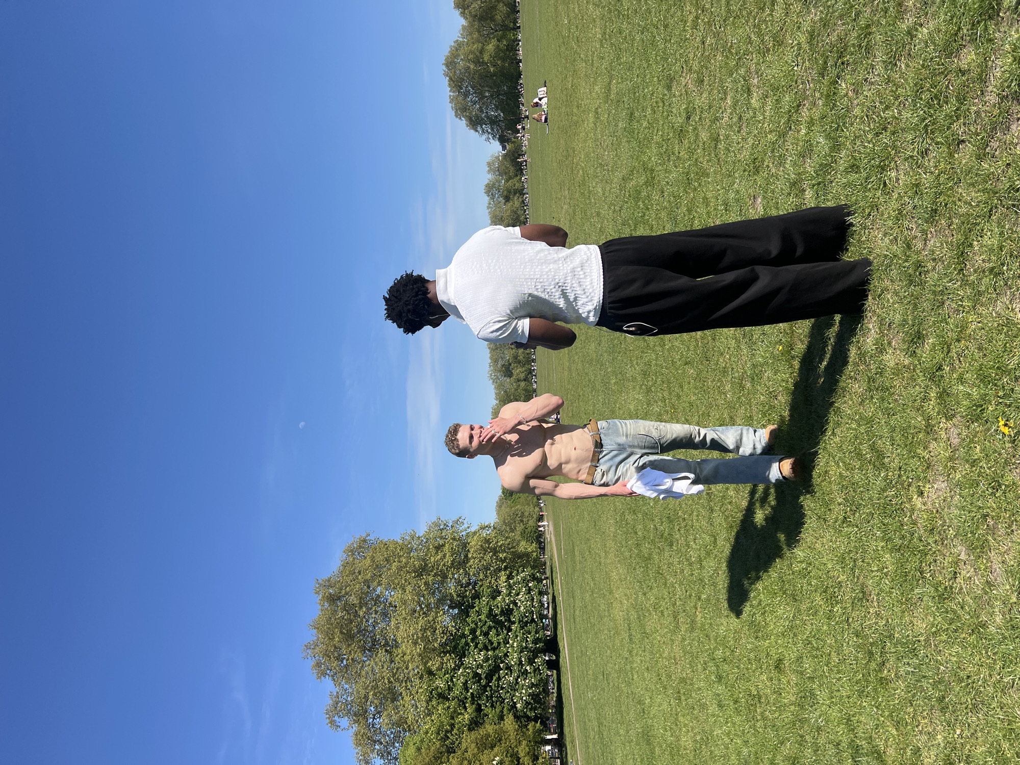 The image shows two men in a park on a sunny day. One man, shirtless, is wearing light-colored jeans and a belt. He is holding a white shirt and has a silver chain around his neck. The other man is wearing a white shirt and black pants. They are standing on a grassy area, and trees are visible in the background. Other people are visible on the grass in the background. The sky is blue with a small moon at the top left corner. The location is likely a park in London, United Kingdom.