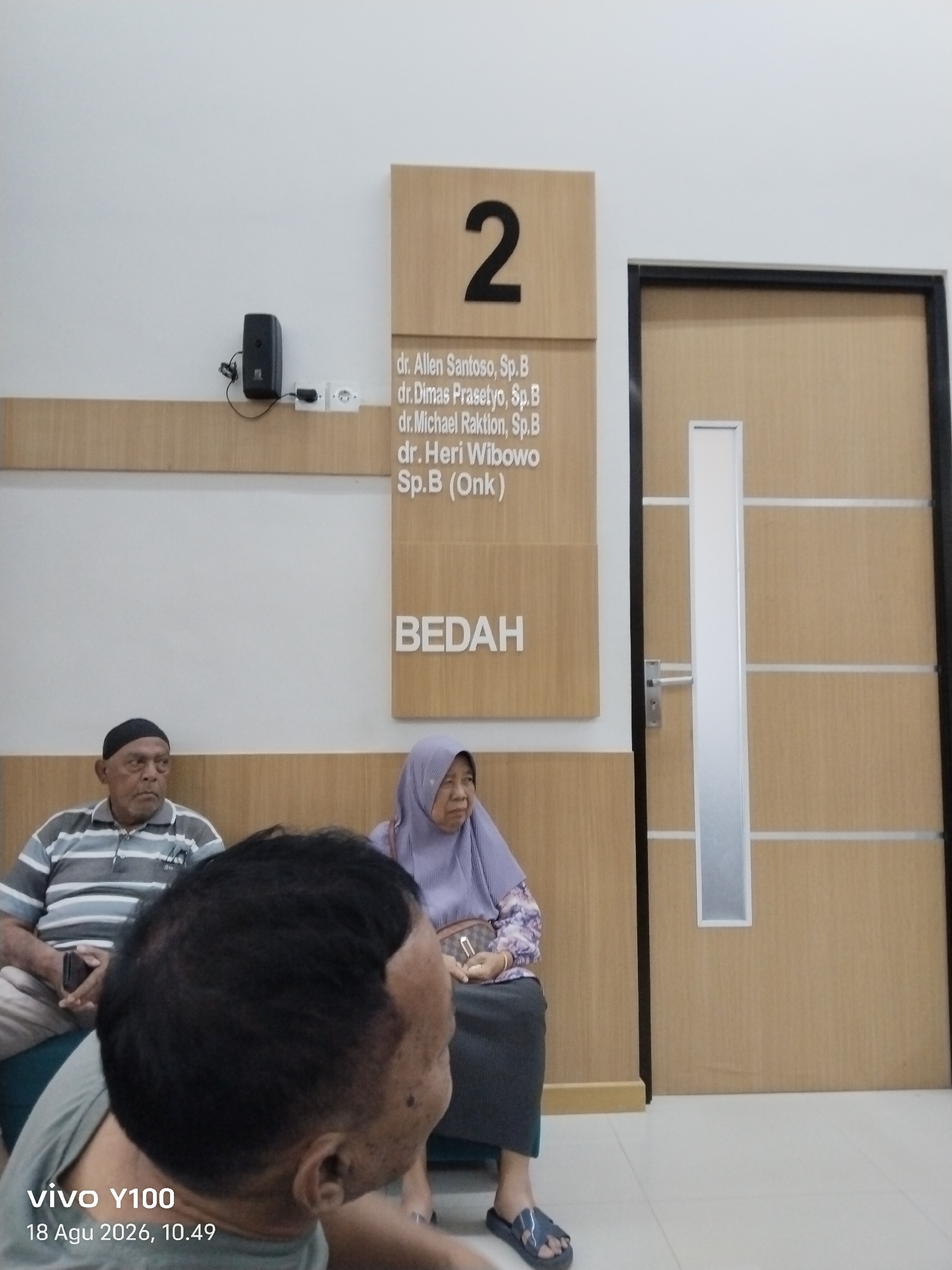 The image depicts a waiting area, likely within a medical facility in Patokan, Indonesia. In the foreground, a person's head and shoulder are visible, facing away from the camera. To the left, an elderly man wearing a striped polo shirt and a black skullcap sits, holding a mobile phone. To his right, a woman wearing a purple hijab and a patterned blouse is seated. Behind them, a door with vertical paneling and a silver handle is visible. Mounted on the wall above the seated individuals is a wooden sign displaying the number "2" and a list of names, including "dr. Allen Santoso, Sp.B" and "dr. Heri Wibowo Sp.B (Onk)". Another wooden sign below this reads "BEDAH". A black speaker is mounted on the wall to the left of the signs, with a white electrical outlet nearby. The floor is tiled with light-colored squares. The overall lighting suggests an indoor environment. The bottom left corner of the image displays a "vivo Y100" watermark and a date and time stamp "18 Agu 2026, 10.49".