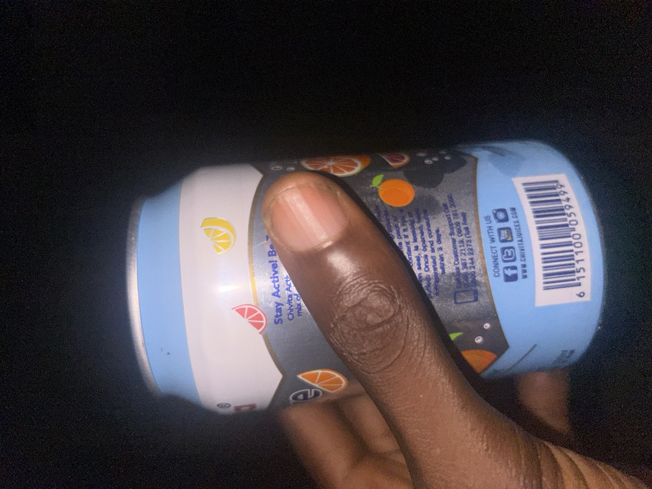 In Egbe, Nigeria, a close-up image depicts a dark-skinned hand holding a Chivita Active beverage can. The can features a light blue and white top and bottom, with a dark grey mid-section displaying illustrations of sliced lemons, grapefruits, and oranges. Prominently visible text on the label includes "Stay Active!", "Chivita Active", customer support telephone numbers (0806 387 2113, 0809 761 2000, 0800 244 2273), social media icons with "CONNECT WITH US", and the website "WWW.CHIVITAJUICES.COM". A UPC-A barcode with the number 6 151100 059499 is visible at the bottom. The individual's index finger wraps across the can's label from the right, with the thumb partially visible on the left side. The background is uniformly dark, indicating low light conditions.
