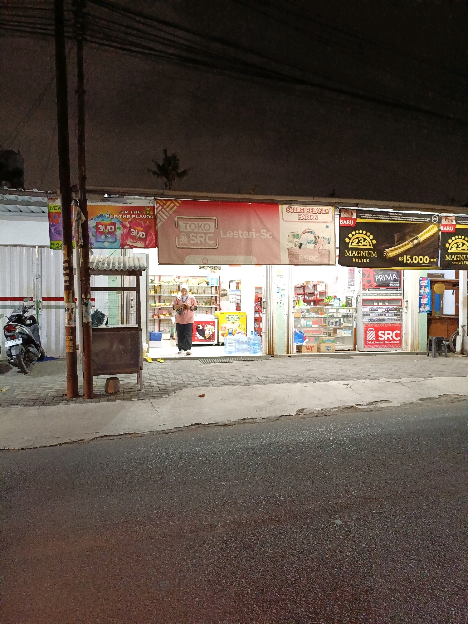 This is a medium shot, taken at night, of a small convenience store or "warung" on a street in Maguwoharjo, Indonesia. The store has a glass-fronted entrance and is lit from within, revealing shelves stocked with various goods. A woman wearing a pink shirt, dark pants, and a headscarf is standing just inside the entrance, looking at her phone. In front of the store, on the sidewalk, is a food cart with a metal exterior and a gray metal roof. A motorcycle is parked to the left of the store. Several signs are visible, including one that reads "Toko SRC Lestari-Sc" and others advertising products like "3VO" and "MAGNUM KRETEK". The street in the foreground is asphalt and appears wet, possibly from recent rain. Overhead power lines are visible against the dark sky.