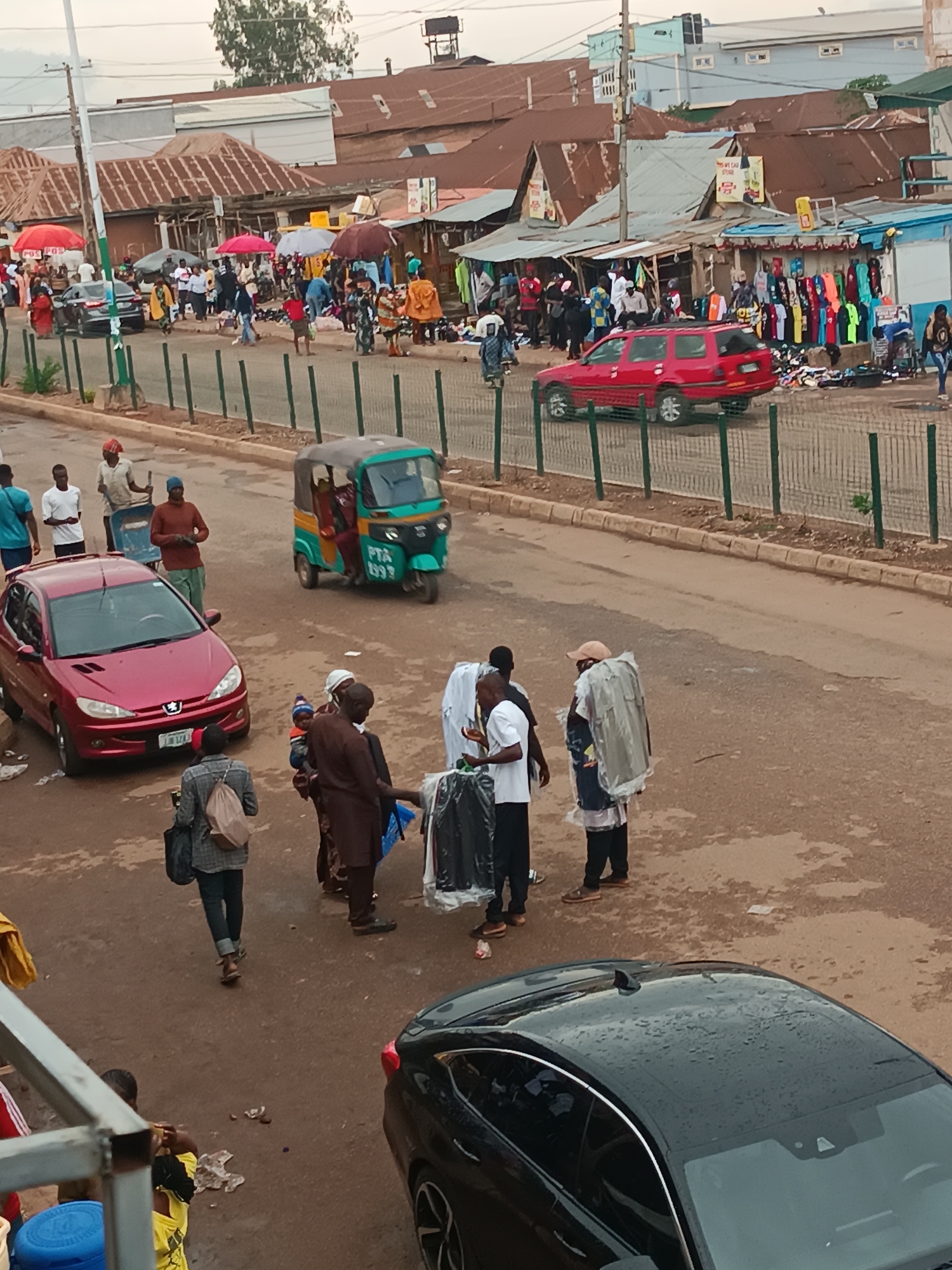 The image depicts a street scene in an urban area. The road surface is reddish-brown. Several people are present on the street, some gathered in groups and others walking. Various vehicles are visible, including a red Peugeot car, a green and yellow auto-rickshaw, and a black car. A market area is located beyond a fence, with stalls displaying goods, including clothing. The buildings in the background appear to have rusty roofs. The sky is overcast. The scene has a bustling, everyday atmosphere, suggestive of a busy street market. The location is in the city of Unknown, Unknown.