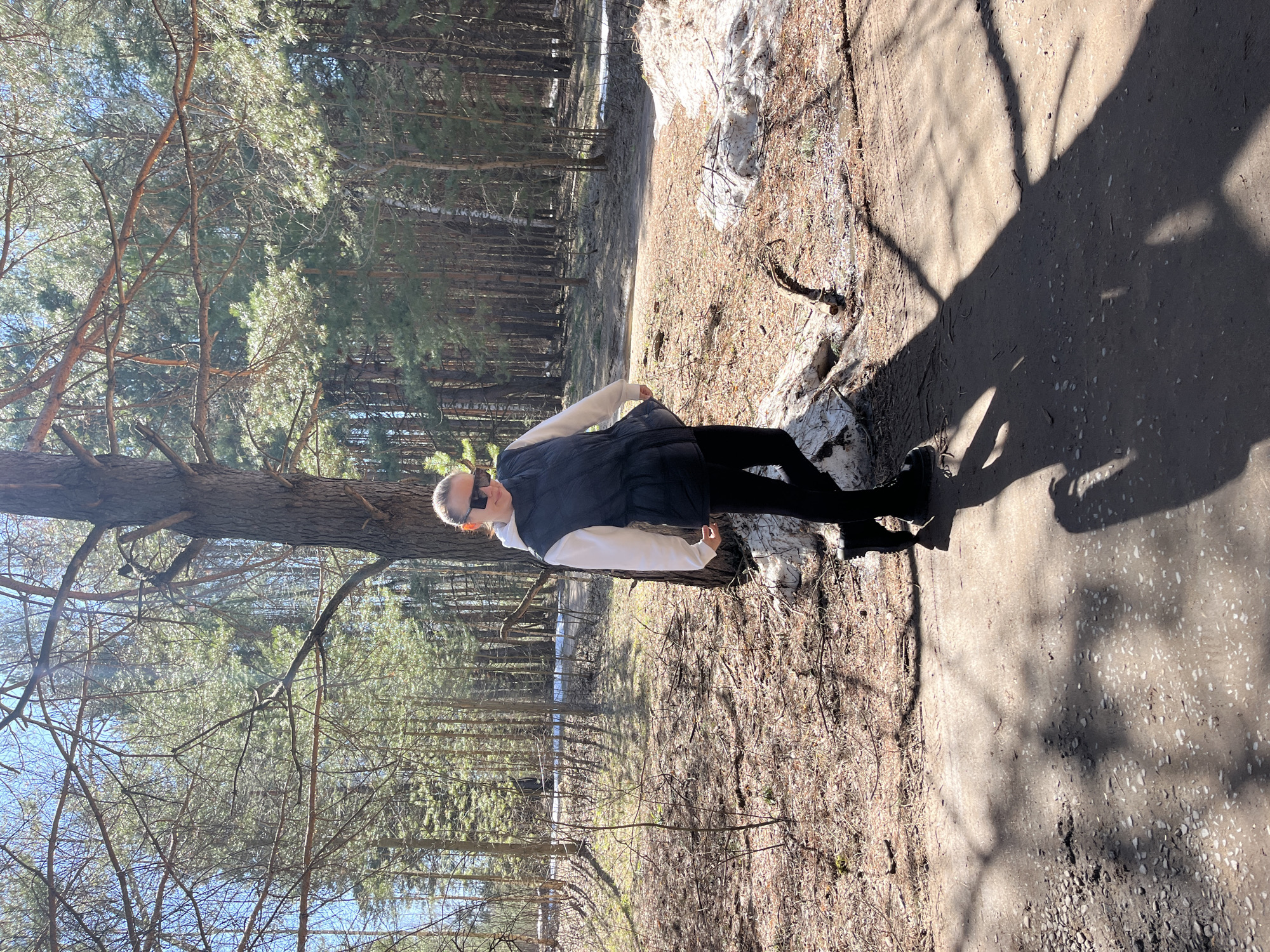 A photograph shows a person standing on a dirt path in a wooded area. The person is wearing sunglasses, a white long-sleeved shirt, a dark vest, and dark pants with boots. The trees in the background are mostly bare, with some evergreen trees visible. Some patches of snow can be seen near the path and among the trees. The location is believed to be in Chaikovsky, Russia.