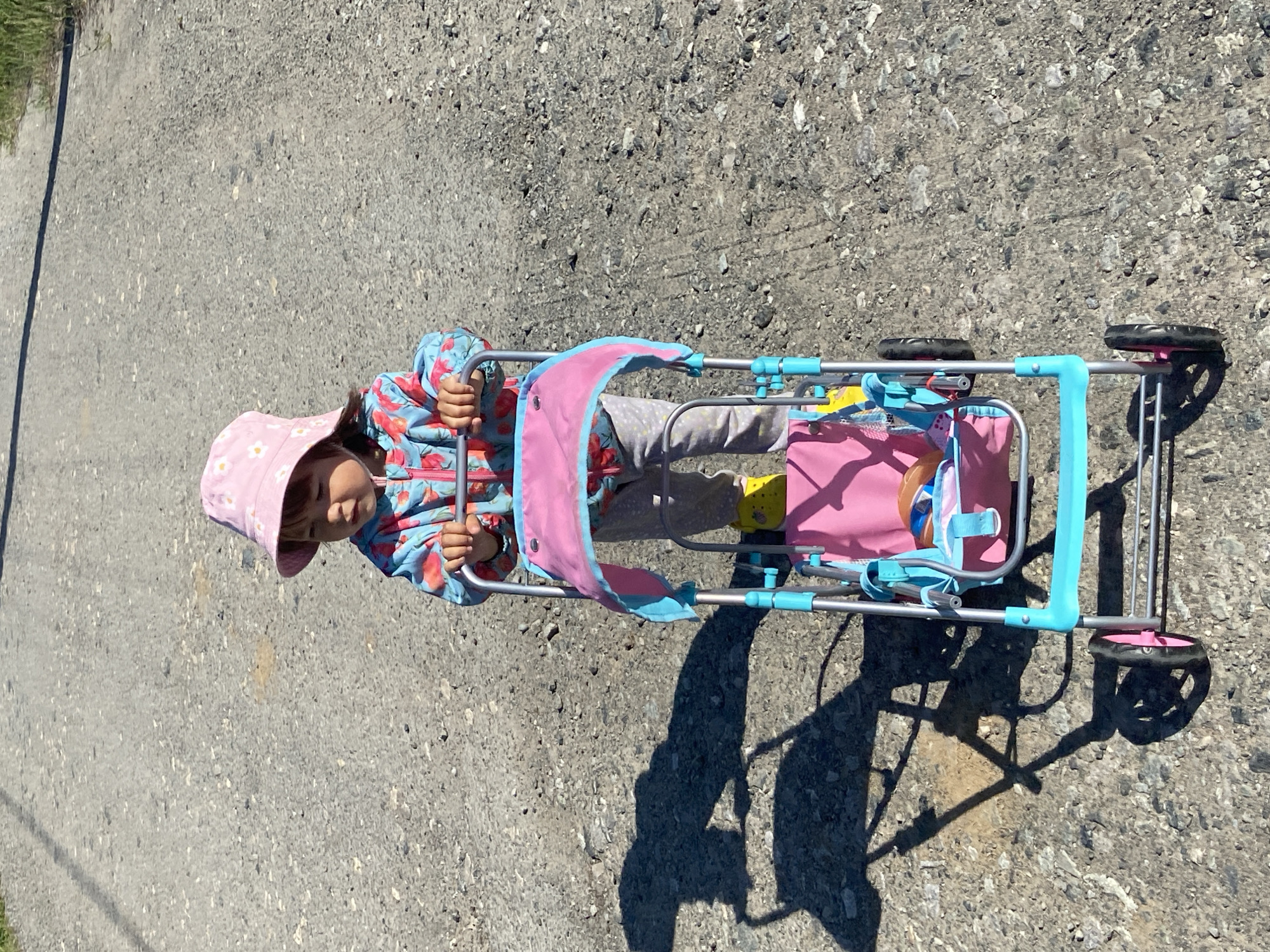 A child, possibly a toddler, is seen lying in a toy stroller on a paved surface. The child is wearing a pink hat and a floral-patterned jacket. The stroller is turquoise and silver with a pink seat and contains a doll. The location appears to be outdoors on a paved surface, possibly a sidewalk or road. There is no definitive information in the image that places it within the city of Tyumen, Russia.