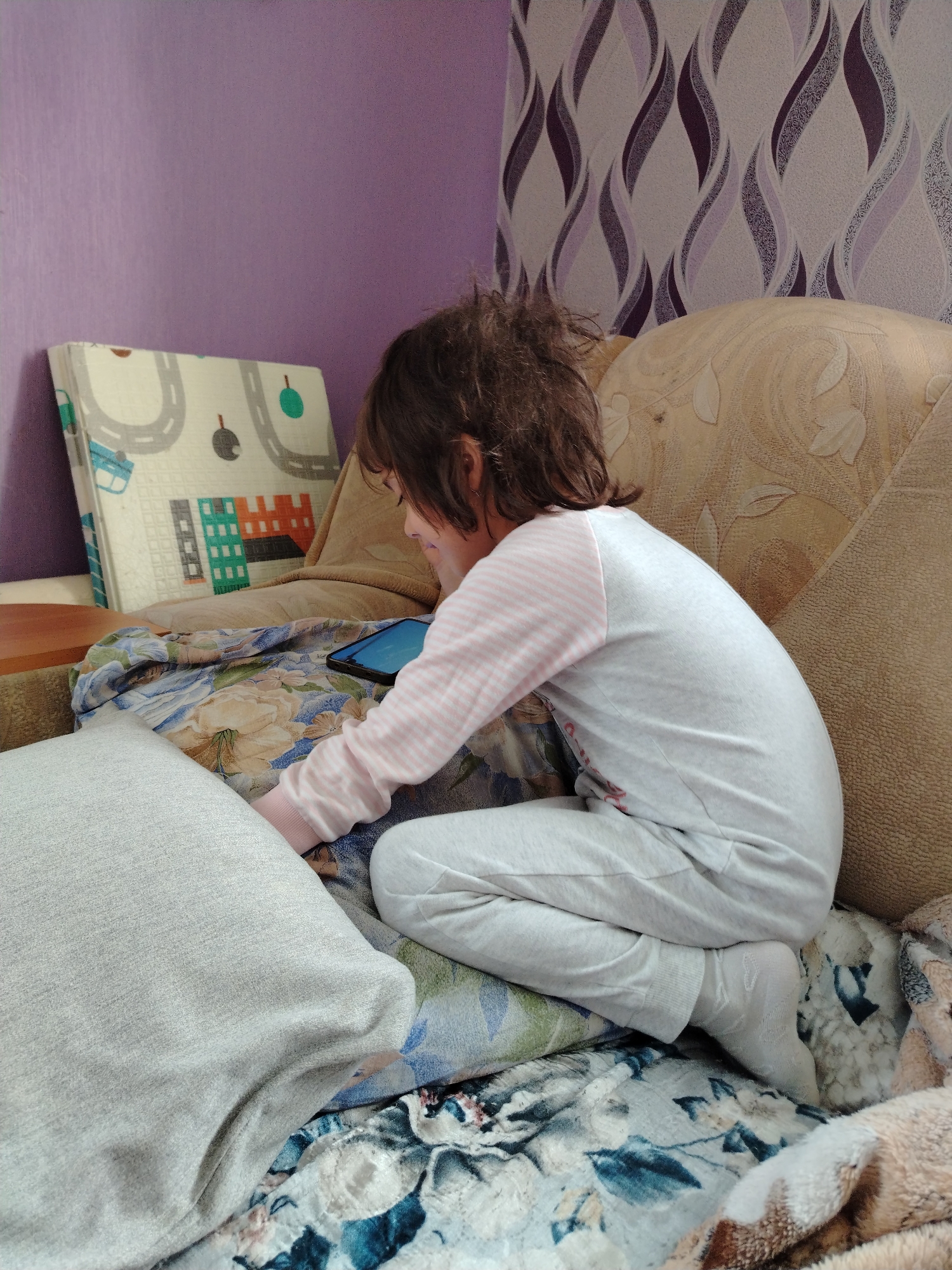 A child with messy brown hair, wearing a light grey pajama set with pink striped sleeves and white socks, is sitting on a sofa, looking at a small black tablet. The sofa is covered in floral patterned blankets and has a large beige patterned back cushion. To the child's left is a grey pillow. Behind the child is a purple wall, and to the right, a wallpaper with a purple and white swirl pattern. Leaning against the wall to the left is a folded mat with a street and building design. The location is assumed to be in the city of Гожан, Russia.