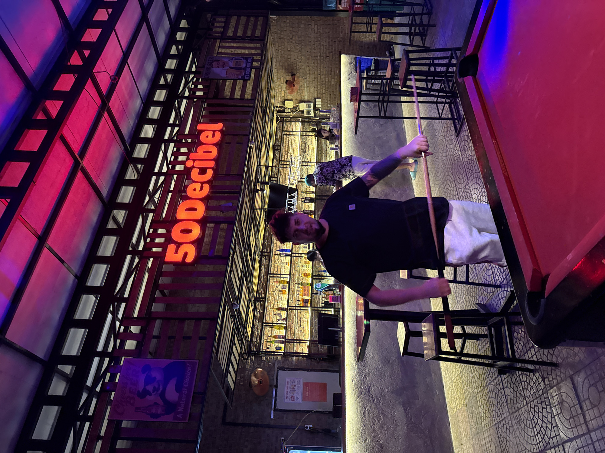 This image captures the interior of "50Decibel," a bar or entertainment venue in Đà Nẵng, Vietnam, likely during the evening or night. The main subject is a man in the foreground, facing the camera, holding a pool cue. He has a beard, is wearing a black t-shirt and white shorts, and displays tattoos on his arm. He stands next to a red-felt pool table, which is illuminated by a blue light reflection.
The venue features an industrial design with exposed brick walls and a structure of wooden planks and metal beams across the ceiling and upper walls. A prominent orange neon sign spells out "50Decibel." The lighting is atmospheric, with red and pink lights reflecting from overhead panels and blue light illuminating the pool table.
In the background, a bar area is visible with shelves holding bottles and glasses. Several tables and stools are scattered throughout the space, and another person in a patterned shirt is partially visible. Posters decorate the walls; one reads "DRINK BEER IT'S GOOD FOR YOU" with an image of two men, and another, featuring a panda-like character with beer, states "Cheers for BEER! A Natural Choice!" and "OBEY." An oscillating fan is mounted on a brick wall. The floor is tiled with a repeating circular pattern. The scene conveys a relaxed, social atmosphere typical of a bar.