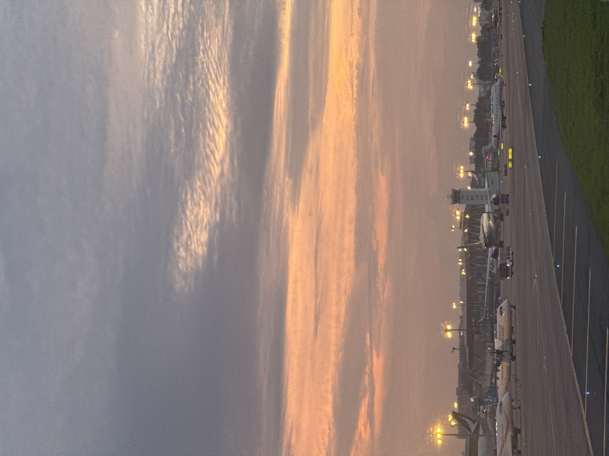 The image depicts an airport tarmac at dusk, with a dramatic sky filled with hues of grey, orange, and pink. Numerous aircraft, including commercial jets and smaller planes, are visible on the ground, some with lights illuminated. A control tower stands tall in the background, surrounded by airport buildings and lights. The scene captures the ambiance of an active airport during the transition from day to night, likely within or near Tan Son Nhat International Airport (SGN), the primary airport serving Ho Chi Minh City, Vietnam, given the characteristic architectural elements and the presence of various aircraft types common to the region. The overall atmosphere is serene yet bustling, indicative of air travel operations.