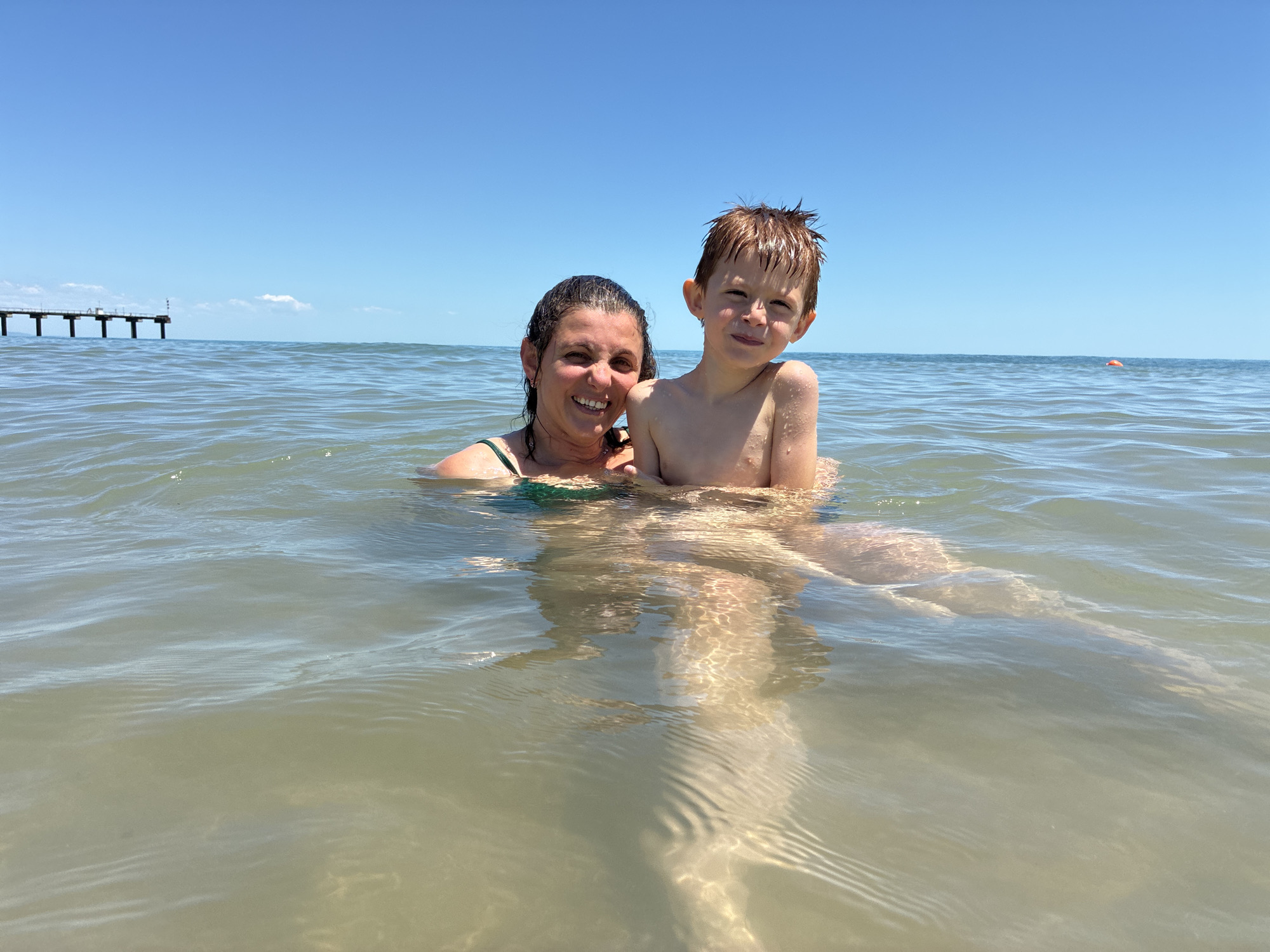 In Shkorpilovtsi, Bulgaria, a woman and a young boy are in the shallow, clear sea under a bright blue sky. The woman, wearing a dark green swimsuit, is partially submerged, smiling and looking at the camera. The boy, with sandy blonde hair, is positioned in front of her and also faces the camera with a slight smile. Sunlight filters through the water, illuminating their bodies and the seabed. In the distance, a pier with pilings extends into the sea on the left, and a small orange buoy is visible on the right. The water is calm, with gentle ripples on the surface.