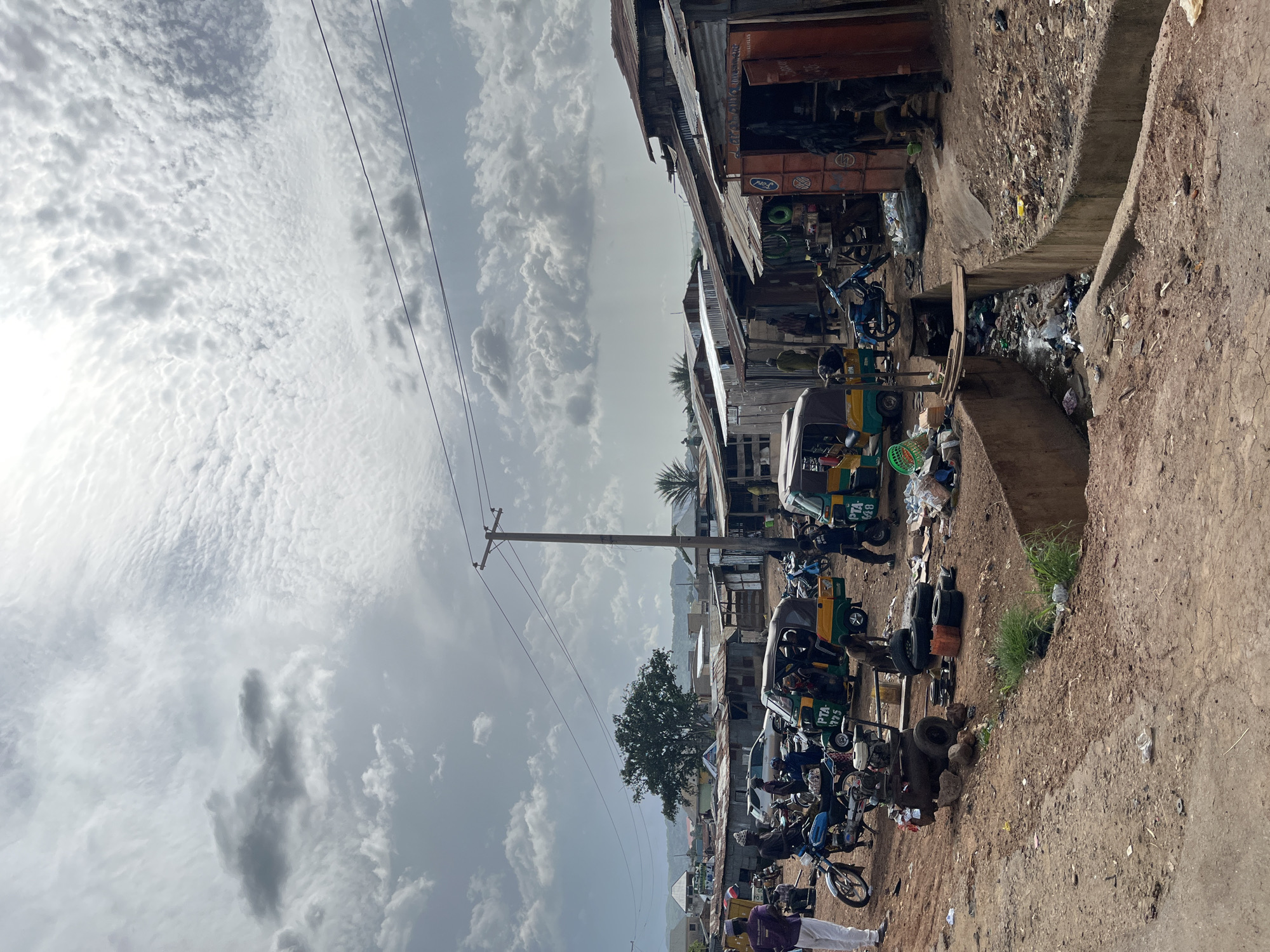 In Jos, Nigeria, a bustling street scene unfolds under a cloudy sky. Several auto-rickshaws, known locally as "keke napep," are parked or in motion, suggesting a busy transportation hub. A man in white pants and a purple shirt stands near a motorcycle, while others mill about the vehicles and the roadside shops. The shops themselves are constructed from corrugated metal, typical of informal settlements or markets. A drainage ditch runs through the scene, filled with refuse, highlighting the environmental conditions. Power lines crisscross the sky, indicating the presence of infrastructure. The overall impression is one of daily life and commerce in a vibrant, albeit somewhat disorganized, urban environment.