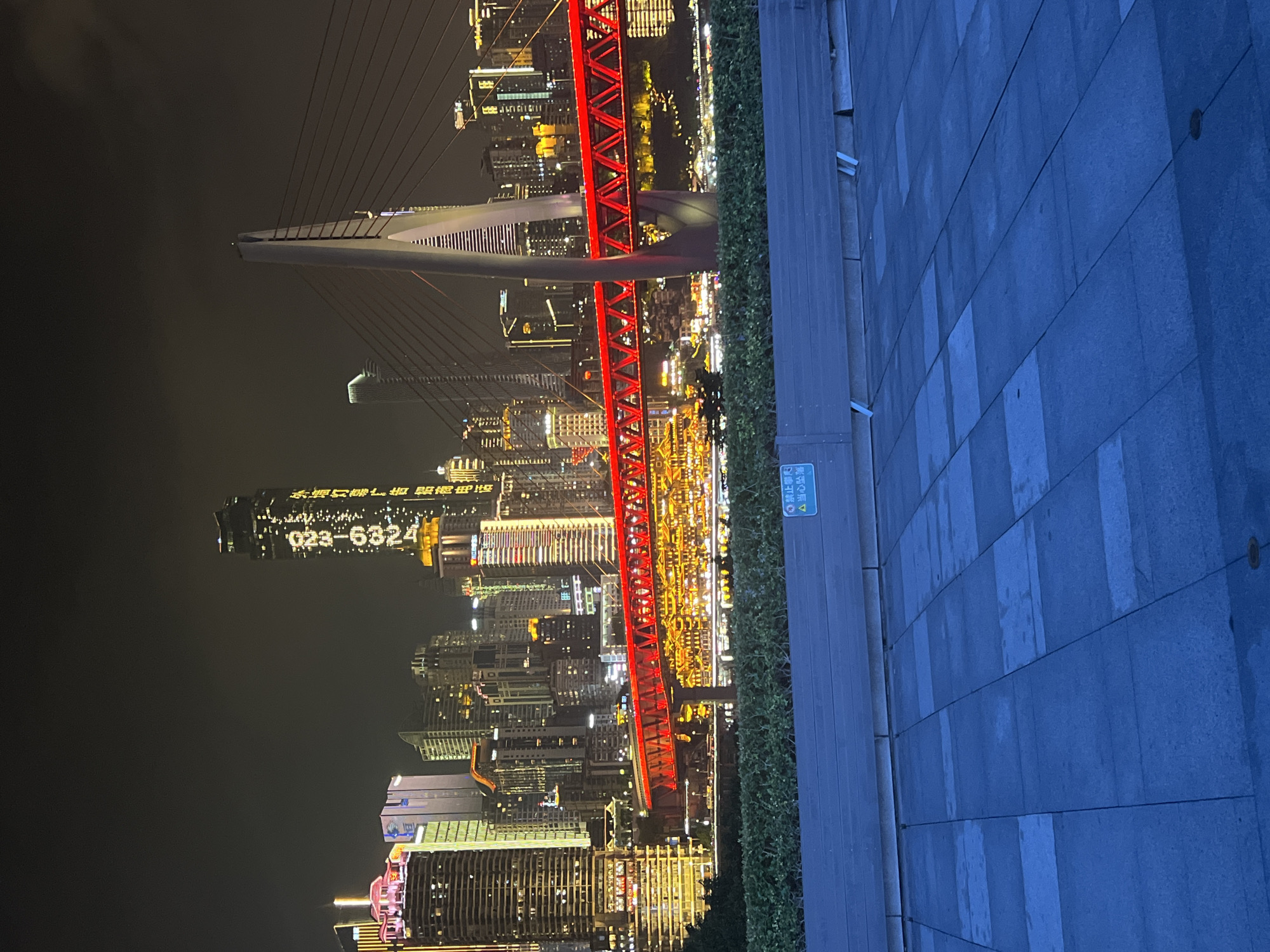 The image captures a night view of the Jiangbeicheng Street in Chongqing, China, showcasing a vibrant cityscape dominated by illuminated skyscrapers and a striking red bridge. The foreground features a dark blue tiled pavement and a low wooden barrier with lush green bushes behind it. A sign with Chinese characters and a warning symbol is visible on the barrier. The middle ground is dominated by the red Hongyadong Bridge, its lights reflecting on the water below. The background presents a dense cluster of modern high-rise buildings, many of which are lit up with colorful lights, creating a dazzling urban panorama. A particularly tall skyscraper in the center displays a large digital readout, possibly indicating a date or time, with "2023" prominently visible. The sky is dark, suggesting nighttime.