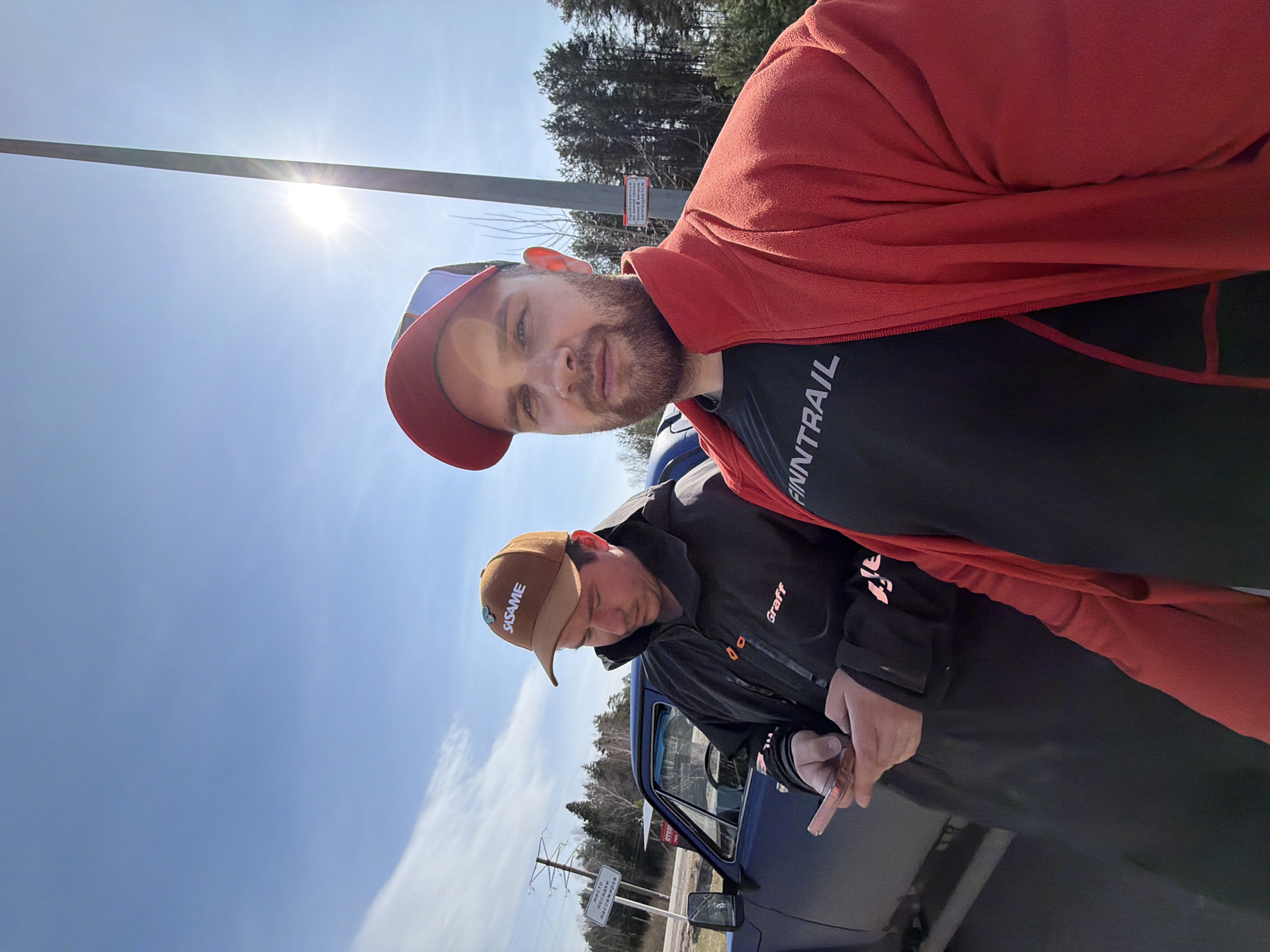 The image, set in Chaikovsky, Russia, shows two men outdoors on a very bright, sunny day with a clear blue sky. The sun is prominently visible in the upper left, creating a starburst effect.
In the foreground, a man with a beard and a neutral expression looks directly at the camera. He wears a red and blue baseball cap, a black t-shirt with "N TRAIL" visible (likely "FINNTRAIL"), and a red fleece jacket draped over his shoulders.
Behind him and slightly to the right, a second man stands next to a blue vehicle, possibly a pickup truck or older SUV, whose driver's side door is open. This man is looking down, wearing a brown baseball cap with "SASAME" on it and a black jacket with "Graff" visible on the sleeve. He holds a slender, cylindrical object, possibly a cigarette or vape pen, in his right hand.
The background features tall trees, power lines, and a utility pole with a sign. The sign, written in Cyrillic, displays warnings such as "ОСТОРОЖНО ПОЛЕВЫЕ РАБОТЫ" (Caution: Field Work) and "проезд ЗАКРЫТ для ПОСТОРОННИХ ПАССАЖИРОВ" (Passage is CLOSED for UNAUTHORIZED PERSONS/PASSENGERS). The scene suggests a casual stop by the side of a road or in a parking area within a semi-rural setting.