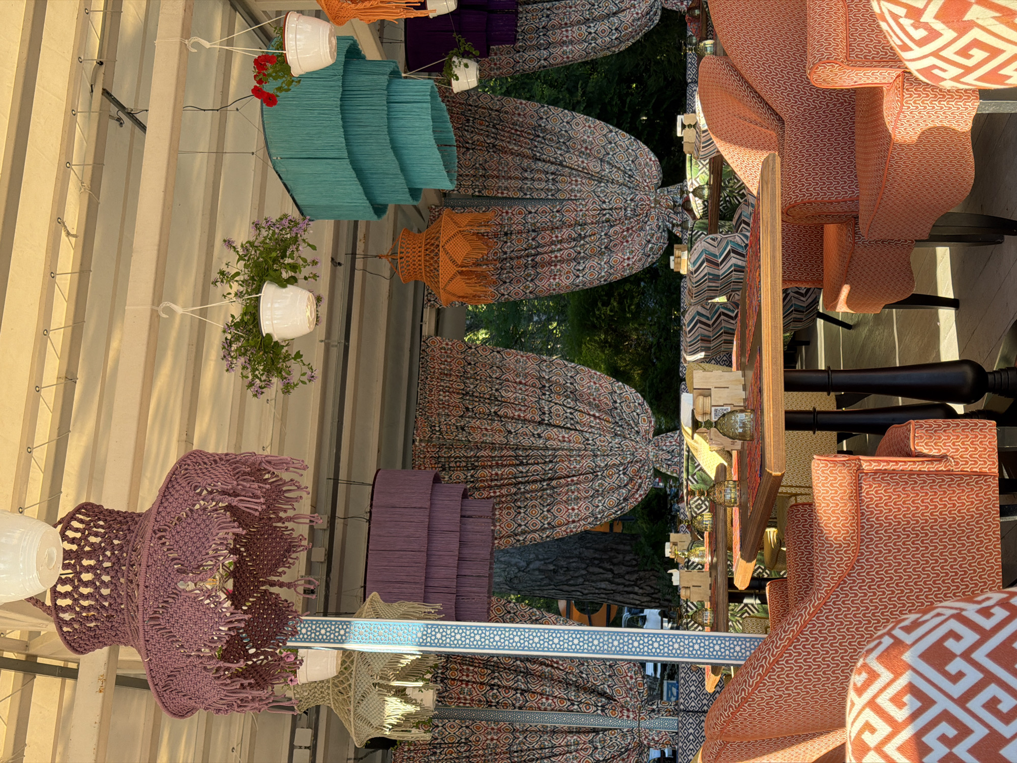This image depicts an outdoor seating area, likely a cafe or restaurant, in Komarovo, Russia. The scene is characterized by a vibrant and eclectic decor, featuring numerous hanging decorative elements that resemble macrame and fringed lampshades in shades of purple, teal, and orange. White planters with flowers are suspended from the ceiling structure, which appears to be a covered patio or pergola.
The seating arrangements include several wooden tables with dark bases, set with placemats and glassware. The chairs and banquettes are upholstered in bold, patterned fabrics, predominantly orange with geometric designs and other colorful abstract patterns. In the foreground, two armchairs with an orange and white geometric pattern are prominently featured. Behind the tables and chairs, draped curtains with a detailed, ethnic-inspired print add to the bohemian ambiance.
The background reveals a lush green outdoor environment with trees, suggesting the location is situated amidst nature. The lighting suggests it is daytime, with sunlight casting shadows and highlighting the textures of the fabrics and decorative items. No people are visible in the image. The overall impression is one of a whimsical and inviting outdoor dining space.
