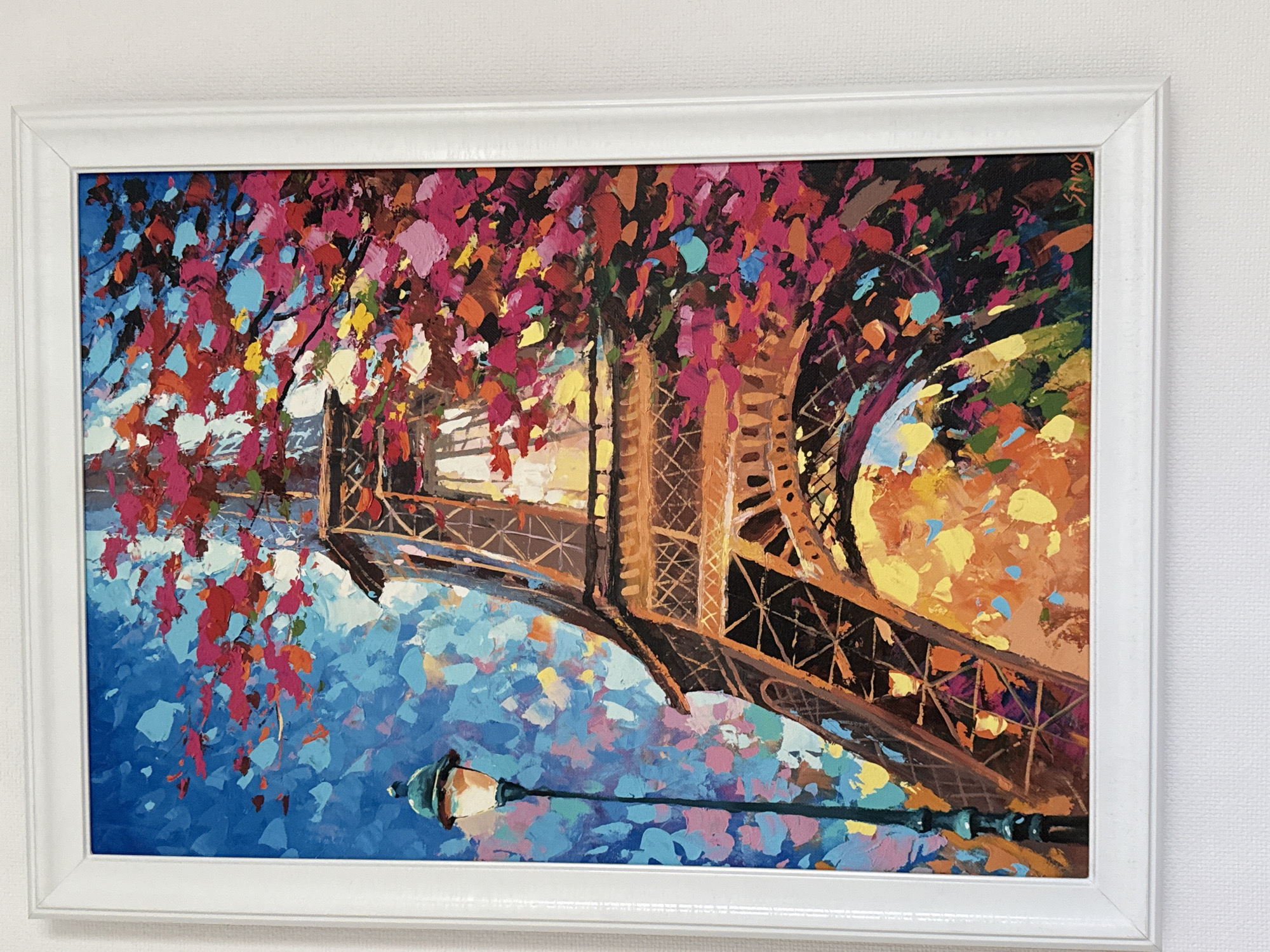 The image is a framed painting of the Eiffel Tower in Paris. The style is impasto, with thick, visible brushstrokes in vibrant colors. The Eiffel Tower dominates the center of the frame, rendered in shades of brown and orange, with glowing yellow light emanating from its base. Surrounding the tower is a dense canopy of foliage depicted in bright reds, pinks, blues, and yellows, suggesting a lively and perhaps autumnal scene. To the left of the tower, a lamppost with a glowing orange light stands against a backdrop of a bright blue sky dotted with colorful abstract shapes. The painting is signed in the lower right corner with the name "SHROS." The framing is simple and white. The painting is hung on a textured white wall. There are no people or animals present in the painting. The location in Burgas, Bulgaria is not depicted in the image.