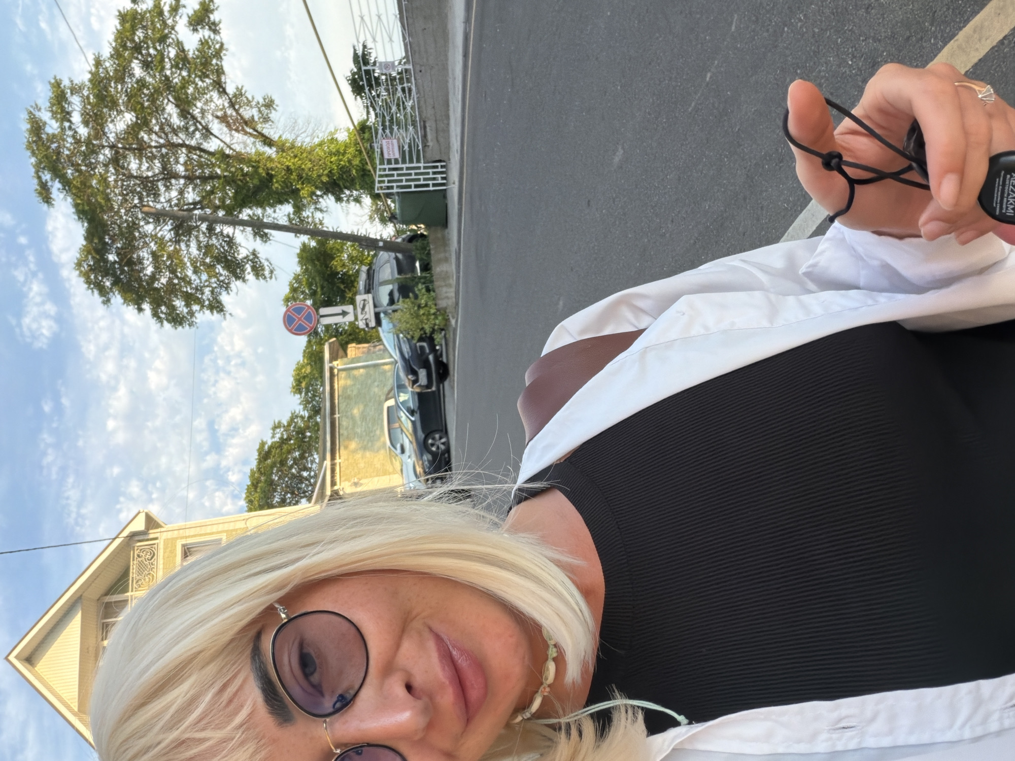 A selfie taken outdoors in Sochi, Russia, shows a woman with blonde hair and glasses looking at the camera. She is wearing a black ribbed top and a white open shirt, with a necklace. In the background, there is a building, trees, cars, a street, and a fence. The sky is partly cloudy.