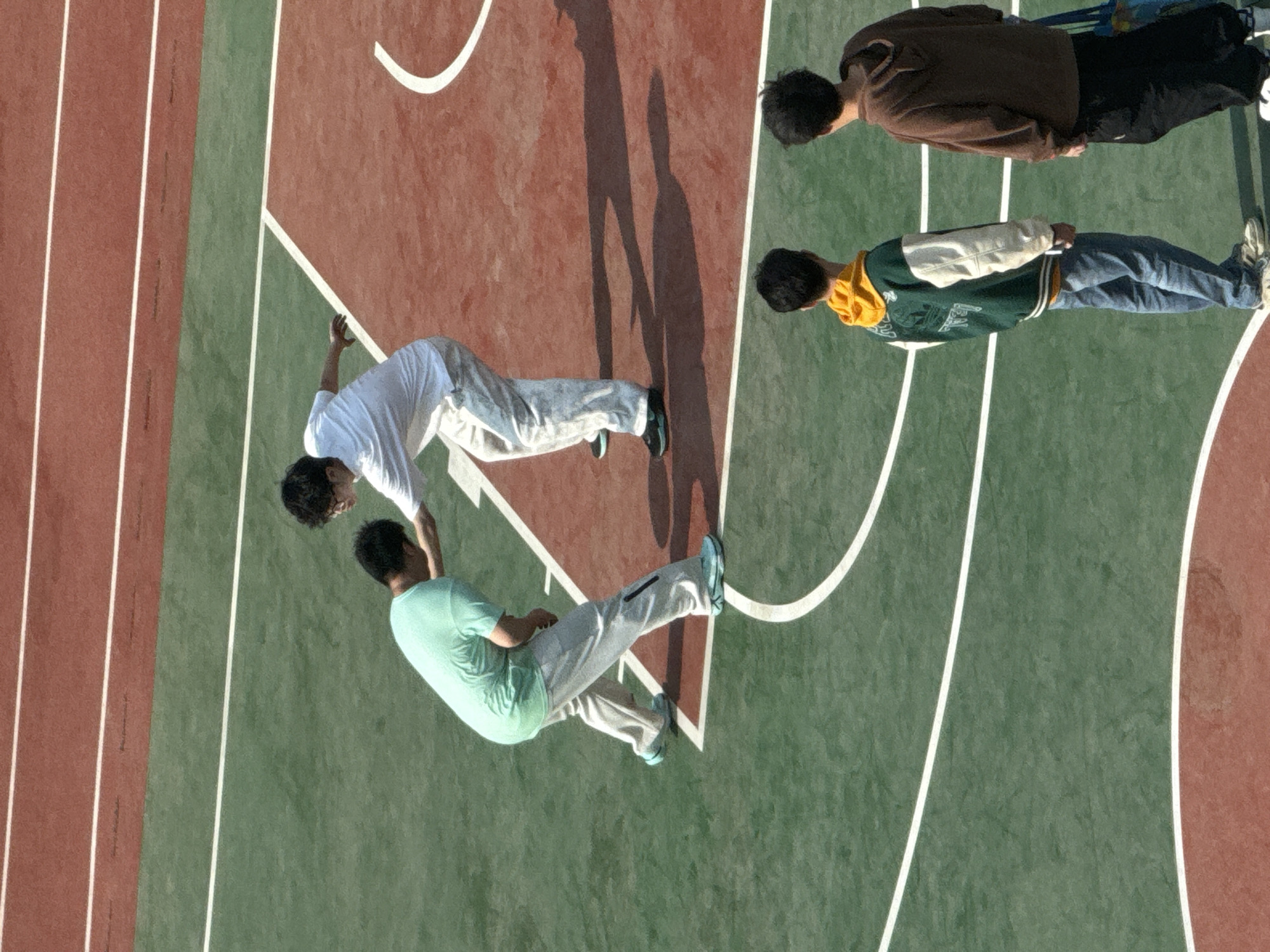 The image presents an overhead view of a multi-purpose sports surface located in the Daxing District, China, containing a running track on the left side and a basketball court on the right. The running track consists of a brick-red surface with white lane markings. The basketball court features a green and brick-red painted surface divided by white lines. The court is unoccupied except for a group of four people who appear to be interacting.
Two individuals closest to the running track are positioned at the edge of the basketball court, one wearing a light mint-green t-shirt and grey sweatpants, and the other wearing a white t-shirt and grey sweatpants. Both people are in a position that appears to suggest they are either falling or leaning. Two other individuals are located on the opposite side of the court, one wearing a brown sweatshirt and black pants, and the other wearing a green and yellow varsity jacket and blue jeans. The shadows of two people indicate that the photo may have been taken during the day with relatively bright sunlight.
