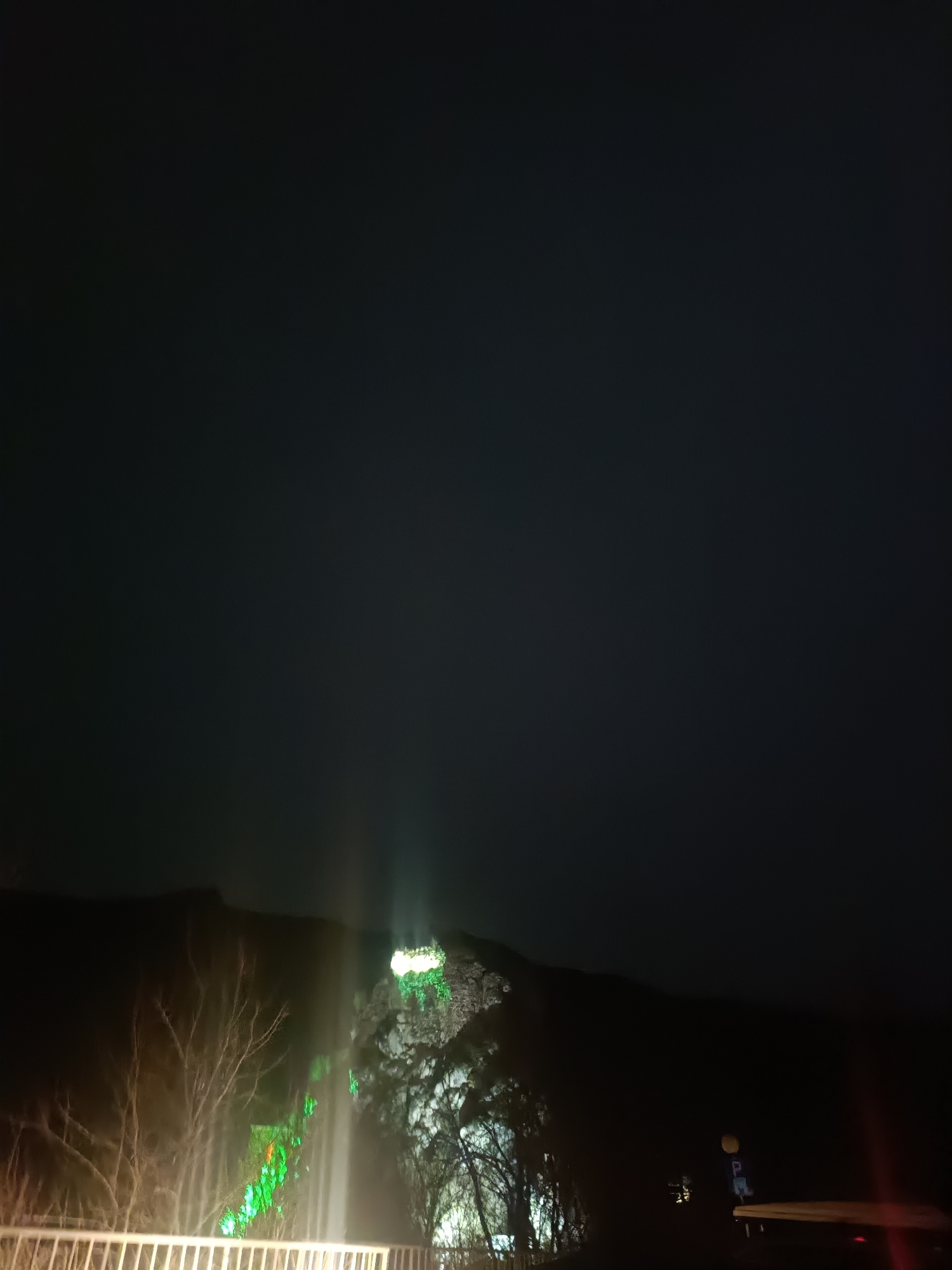 At night, the image shows a hill or mountain illuminated with green lights in the city of Asenovgrad, Bulgaria. The lights appear to be concentrated at the top and partially down the hillside. There is a waterfall or a streak of light in the foreground, near some trees. A white barrier or fence is visible at the bottom of the image. The sky is very dark.