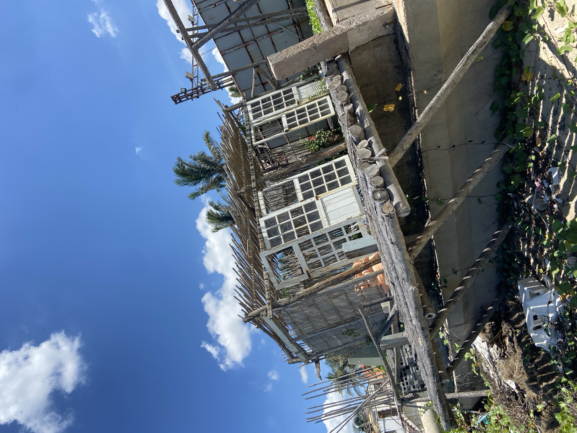 The image shows a dilapidated wooden structure on a sandy beach under a bright blue sky with scattered clouds. The structure appears to be an unfinished or abandoned building, with exposed wooden beams, bamboo roofing, and several white doors with glass panes. The structure is leaning precariously, suggesting structural instability. A raised wooden walkway with a makeshift railing leads up to the structure. The foreground is cluttered with debris, including plastic waste and dried vegetation, with green vines growing around the base of the structure and on the beach. In the background, palm trees are visible, along with other buildings and parts of what appear to be construction or scaffolding. The scene suggests a coastal environment in Phu Thuy Ward, Vietnam, impacted by weather or neglect, leading to its deteriorated state. There are no people visible in the image.