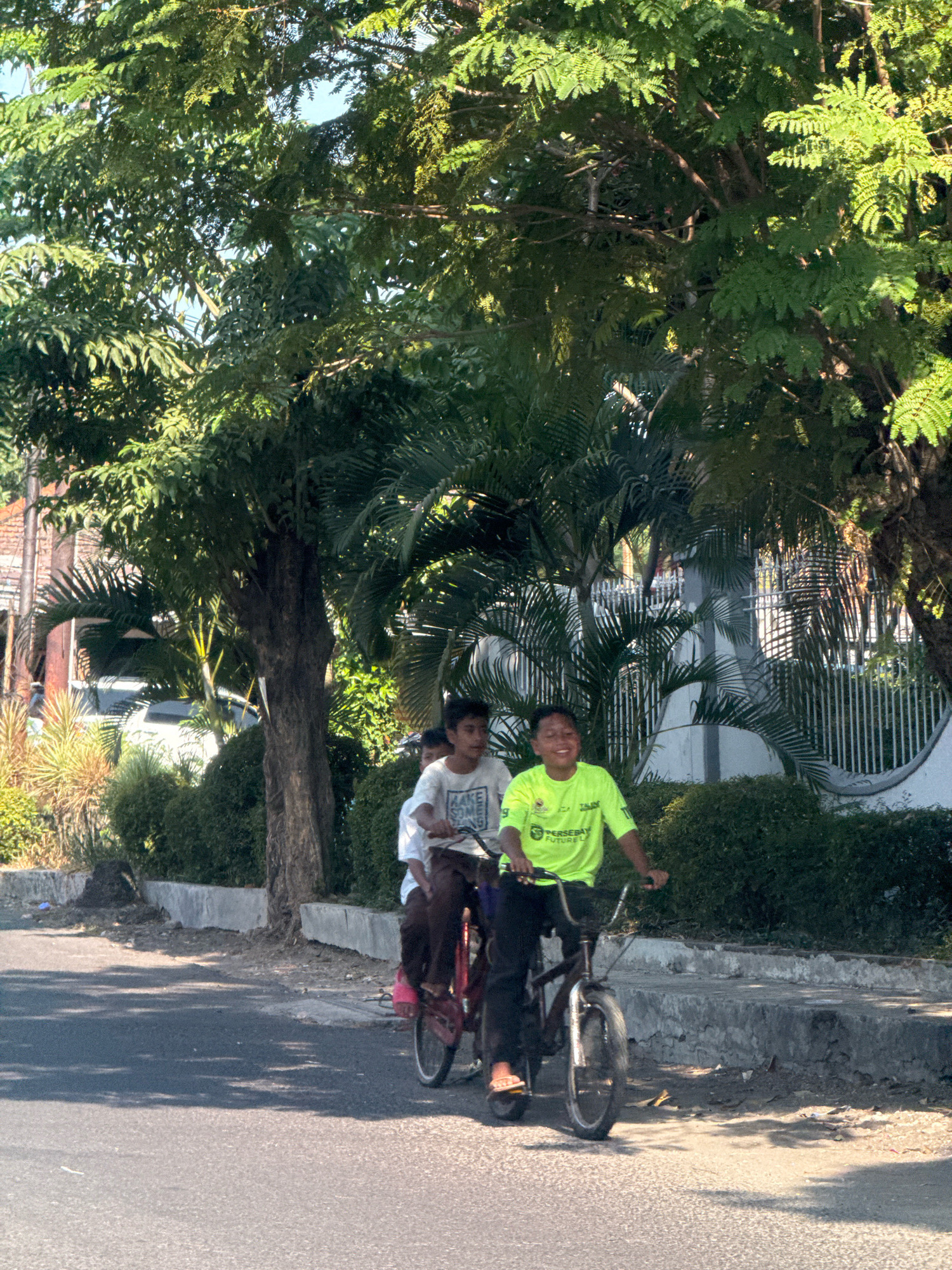 Three young boys are riding on two bicycles on a paved road in Surabaya, Indonesia. The boy in the foreground is wearing a bright green t-shirt with a logo and black pants, and is smiling as he rides his bicycle. He is followed by two boys on a tandem bicycle. The boy in the middle wears a white t-shirt and dark pants, and the boy in the back is mostly obscured by the boy in front. They are riding past a sidewalk lined with green bushes and trees. The sun is shining, casting shadows on the road, and the sky is visible through the leaves of the trees. The buildings in the background are partially visible, and there is a white fence.