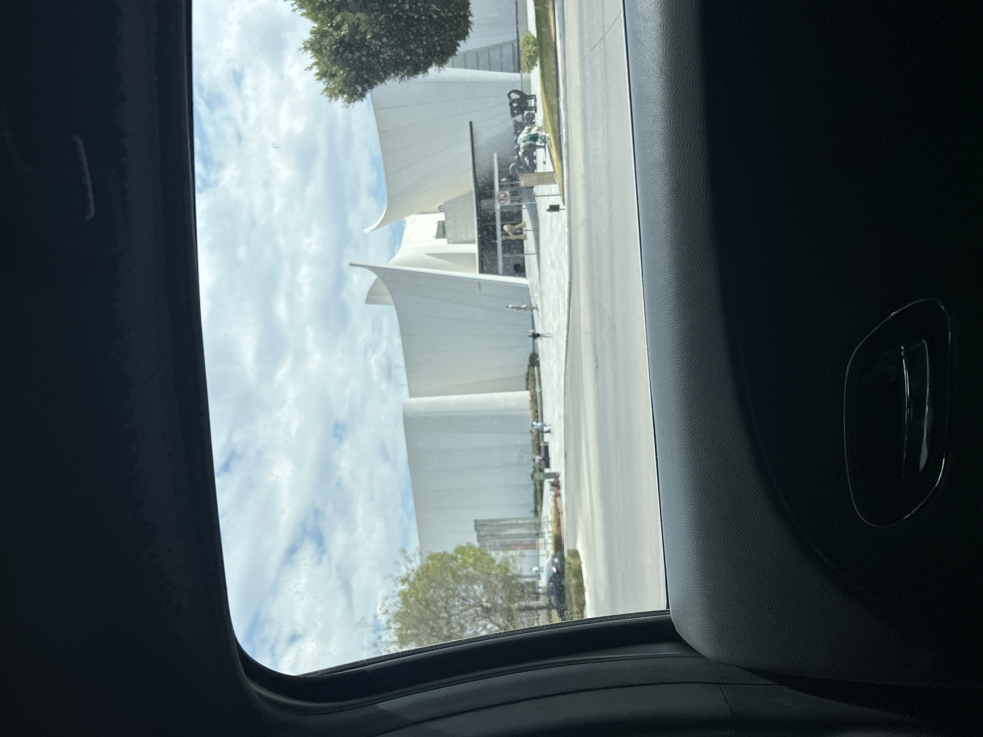 View from inside a vehicle, looking out at a large, white, modern architectural building with swooping, sail-like elements. The sky is cloudy. Other buildings, trees, sidewalks, and parked vehicles are visible outside the building. The photo appears to have been taken while the vehicle was moving.
The building is likely the Museo Internacional del Barroco, located in San Bernardino Tlaxcalancingo, Puebla, Mexico, due to its distinct design.
