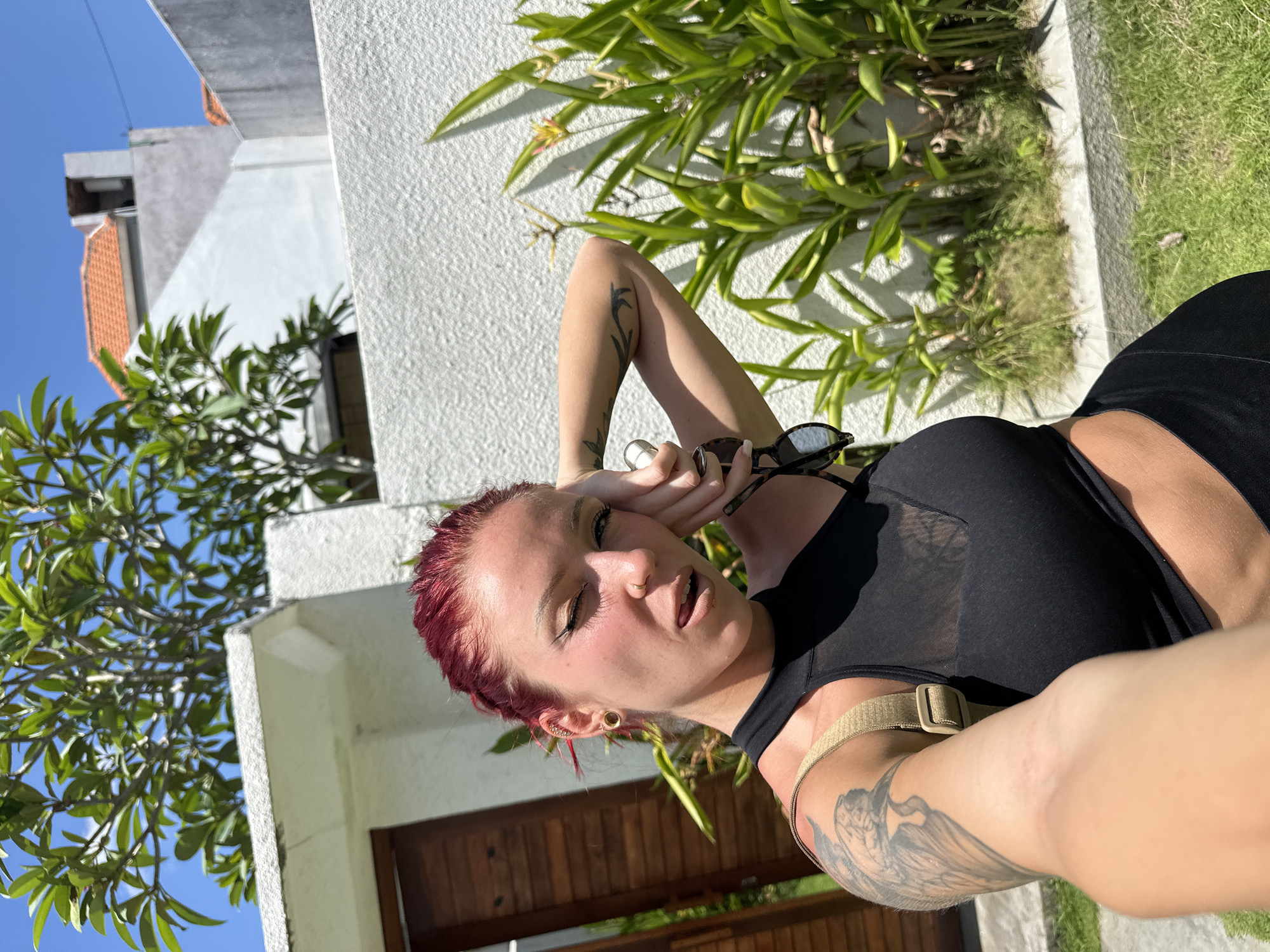 The photograph depicts a woman with red hair lying down in an outdoor setting. She is fair-skinned and has tattoos on her arms. She is wearing a black top with a mesh detail and has sunglasses near her face. The woman appears to be posing for a selfie, with her arm outstretched.
The background features a white wall with green plants growing alongside it. A building with a red-tiled roof and wooden doors is also visible. The scene appears to be located in Dalung, Indonesia, indicated by the architecture and vegetation. The lighting suggests a sunny day.
