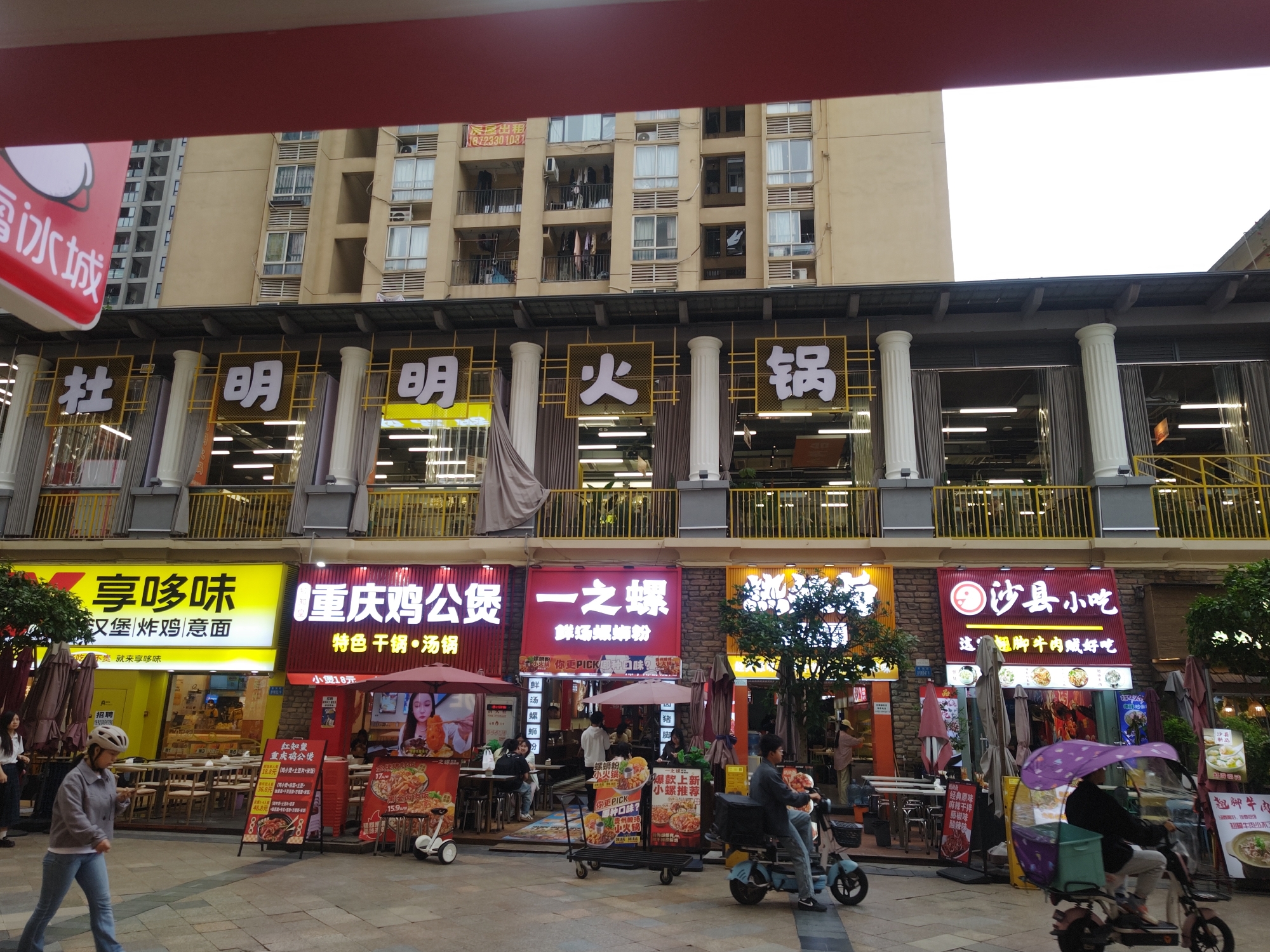 This medium-shot photograph captures a lively commercial street scene in China, featuring a row of restaurants on the ground floor of a multi-story residential building. The upper section of the image shows the beige residential building with numerous windows and balconies, some displaying hanging laundry. A small "for rent" sign with a phone number (18723301031) is visible on a balcony. Below, large white Chinese characters on a yellow background spell out "杜明明火锅" (Du Ming Ming Hotpot), indicating a restaurant on the second level, supported by decorative white columns.
On the ground level, multiple brightly lit eateries line the street, each with its own distinctive signage and some with outdoor seating under umbrellas. From left to right, these include:
* "享哆味" (Xiang Duo Wei), a yellow-fronted shop offering "汉堡 炸鸡 意面" (Hamburger, Fried Chicken, Pasta) and promoting itself with "不贵就来享哆味" (If it's not expensive, come to Xiang Duo Wei), also displaying a "招聘" (Recruiting) sign.
* "重庆鸡公煲" (Chongqing Chicken Pot), a red-fronted restaurant specializing in "特色 干锅·汤锅" (Specialty Dry Pot, Soup Pot), with a "小煲18元" (Small pot 18 yuan) offer and additional signs for "红知皇 重庆鸡公煲" (Hong Zhi Huang Chongqing Chicken Pot) with various prices.
* "一之螺" (Yi Zhi Luo), another red-fronted establishment serving "鲜汤螺蛳粉" (Fresh Soup Luosifen) and "小火锅" (Small Hotpot), inviting customers to "PICK哪种口味?" (Which flavor do you PICK?) from options like "经典原味 麻辣干拌 藤椒味 酸辣味" (Classic original, Spicy dry mixed, Tengjiao flavor, Sour and spicy flavor).
* "沙县小吃" (Shaxian Snacks), also with a red facade, advertising "这翘脚牛肉贼好吃" (This Qiaojiao beef is super delicious) and featuring "翘脚牛肉" (Qiaojiao Beef).
The street itself is paved and bustling with activity. People are walking, including a woman in a grey jacket and helmet on the left. Several individuals are seated at outdoor tables. Various electric vehicles are present, including a white self-balancing scooter and a delivery tricycle with a purple canopy, ridden by a person in the foreground. The time of day appears to be daytime, possibly overcast, given the bright white sky and the illuminated storefronts. A partial sign on the far left reads "冰城" (Ice City).