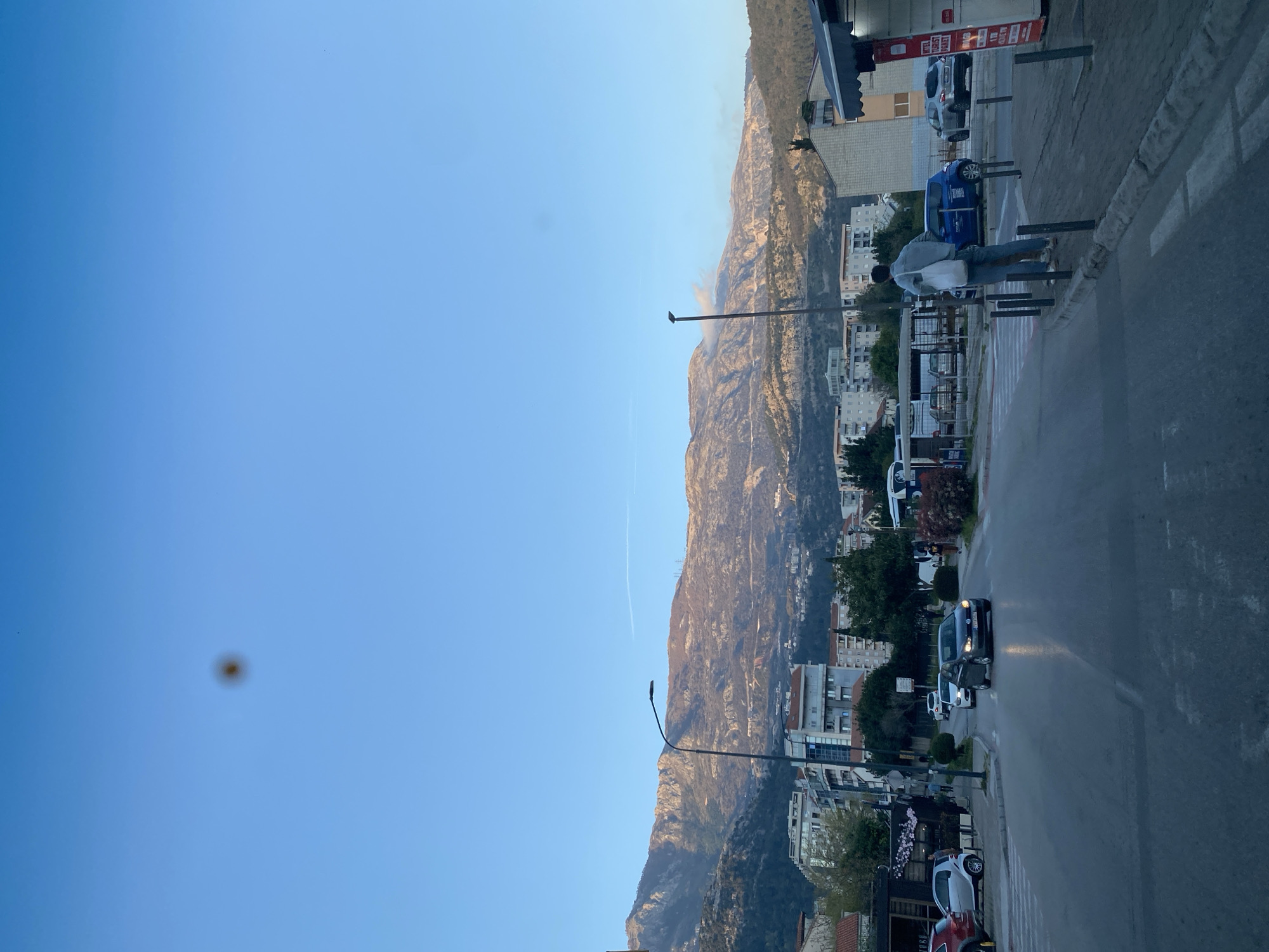 The image shows a street scene in Budva, Montenegro, with a mountain range in the background. The sky is clear blue. There are buildings lining the street, along with parked cars and streetlights. A person is visible standing on the sidewalk near a bus stop. A road runs parallel to the sidewalk. In the far distance, a mountain range is the backdrop, with indications of smoke or haze near the mountains.