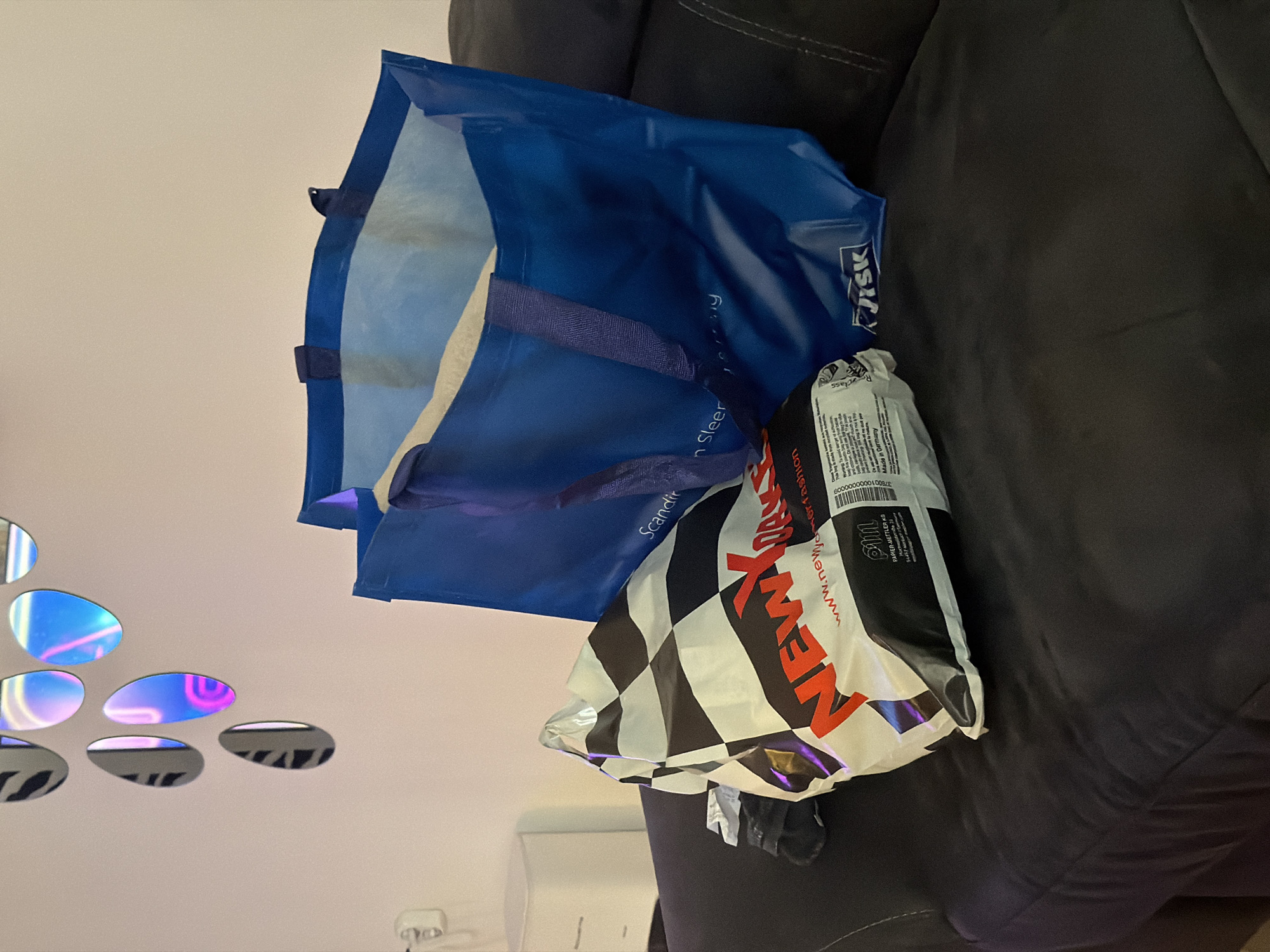 This image captures an indoor, residential scene in Plovdiv, Bulgaria, specifically a portion of a living area. In the foreground, resting on a dark gray upholstered sofa, are two shopping bags. To the left is a white plastic bag featuring a black and white checkered pattern, emblazoned with "NEW YORKER" in prominent red lettering and the website "www.newyorker.fashion". Additional text on this bag includes "PAPIER-BETTLER KG" and "Made in Germany". To the right is a larger, vibrant blue reusable shopping bag with the "JYSK" logo visible at its base, and partial text "Scandinav en Sleep" suggesting "Scandinavian Sleep & Living".
The background consists of a light-colored wall. Adorning the wall are multiple teardrop-shaped decorative mirrors, reflecting iridescent hues of blue, pink, and purple light, creating a dynamic visual effect. To the far left, a white electrical outlet is mounted on the wall. Below the outlet and slightly behind the bags, a small, white, multi-drawer cabinet or nightstand is partially visible. No individuals are present in the frame. The overall impression is a snapshot of items brought home after a shopping excursion, placed casually on a sofa in a contemporary domestic setting.