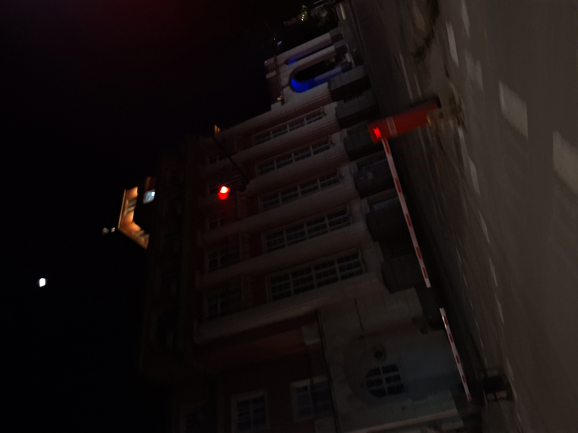 At night, a tall building is viewed from below. The structure appears to be an apartment complex with multiple stories and numerous windows. Some lights are visible within the building. A red signal light is attached to the building on one side, and a safety barrier extends towards a nearby wall. The environment is dark, with a partial moon visible in the upper left. The image location is potentially Pleven, Bulgaria, but there is no visual evidence to support this assertion.