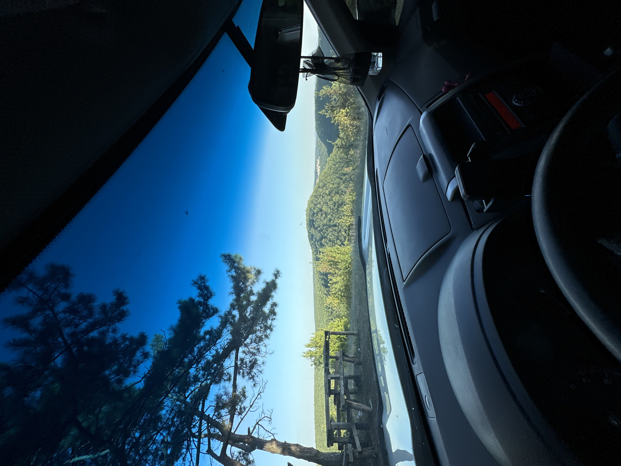 The image depicts a view from inside a car parked in the city of Makiv, Ukraine. Through the windshield, a scenic landscape of rolling green hills covered in dense trees under a clear blue sky is visible. In the foreground, a wooden picnic table and bench are situated beneath the branches of a large tree. The interior of the car, including the dashboard and steering wheel, is also captured. A rear-view mirror hangs from the windshield, and a decorative car air freshener is suspended from it. The overall scene suggests a peaceful outdoor setting, likely a scenic overlook or a rest stop.