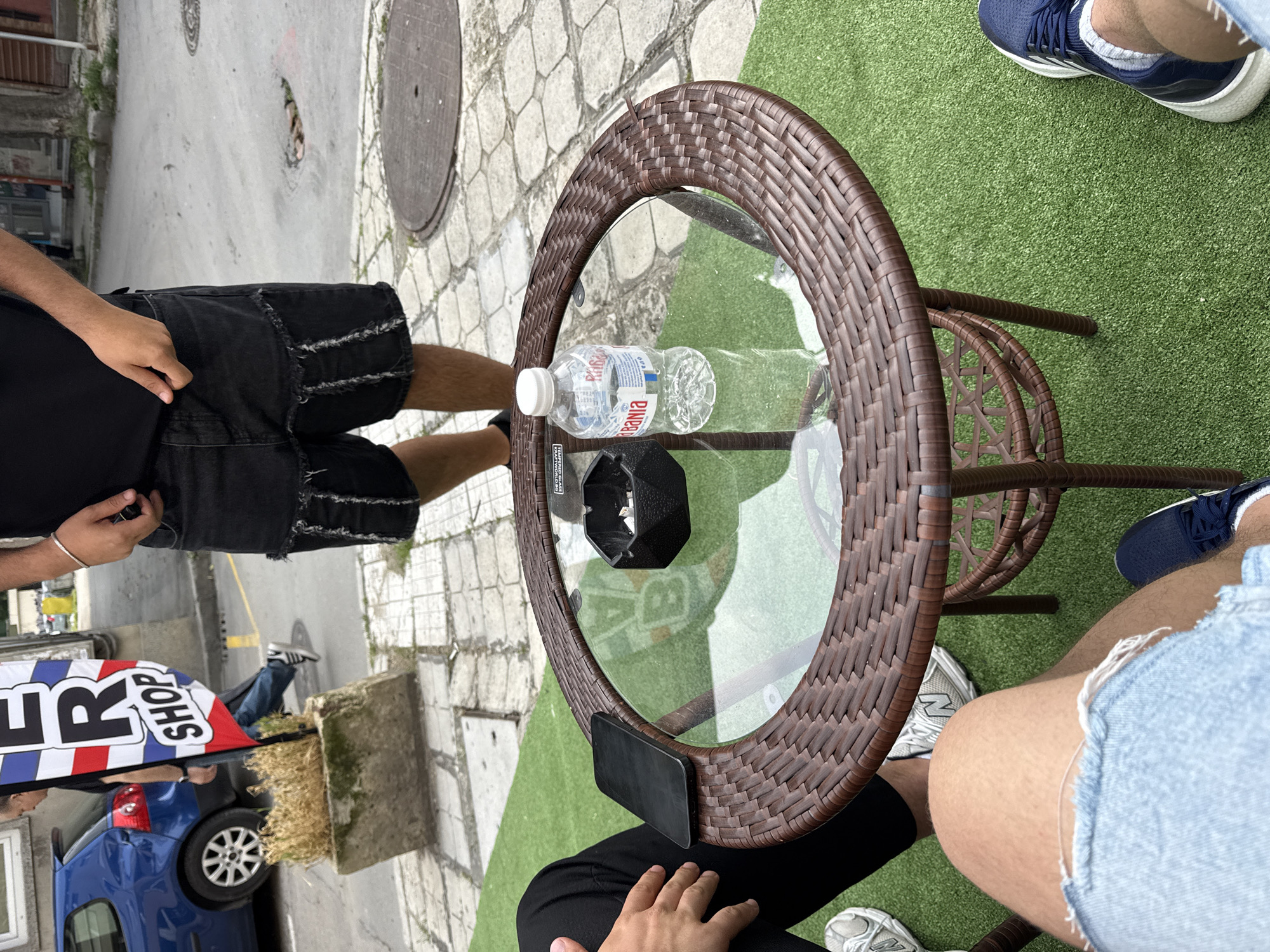 The image captures an outdoor scene, likely a cafe or restaurant seating area, adjacent to a street in Varna, Bulgaria. The perspective looks down upon a round table and the people seated around it.
In the center, a round outdoor table features a glass top and a dark brown, woven frame. On the table, there are two clear plastic water bottles, one of which has a label partially visible with the Cyrillic word "Шипка" (Shipka). A black, hexagonal ashtray and a black smartphone are also present on the table.
Two individuals are seated at the table, their legs and lower torsos visible. The person on the left wears distressed black denim shorts and a black top, with a silver bracelet on their right wrist. The person on the right wears light blue ripped denim shorts and dark blue sneakers with white soles. Their legs are bare. The seating area is covered with artificial green grass.
Beyond the grass, a paved sidewalk with light-colored cobblestones and several manhole covers leads to a street. A blue car is parked on the street. In the background, part of a vertical banner sign is visible, displaying red, white, and blue stripes and the word "SHOP" in white letters, likely indicating a "Barber Shop" or similar establishment. The scene is brightly lit, suggesting it is daytime with clear or partly cloudy weather. The overall atmosphere appears relaxed.