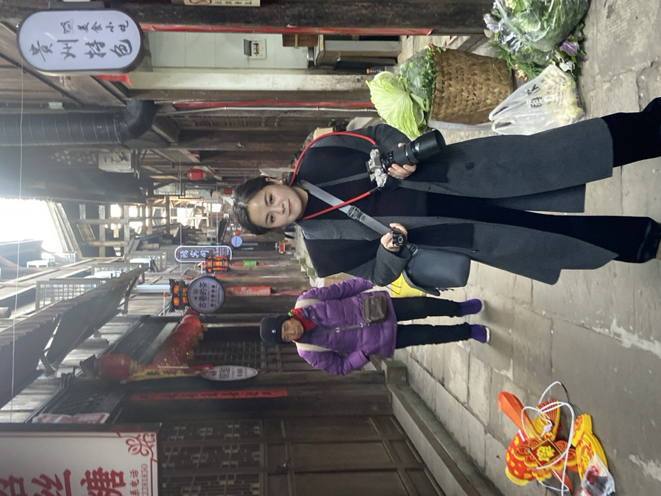 A woman with a camera poses in a narrow, old-fashioned street lined with wooden buildings adorned with signs and red lanterns. Behind her, an older person in a purple coat walks on the cobblestone street. A basket of produce sits near the woman, and a dragon-shaped decoration rests on the ground. The scene suggests a tourist spot or a traditional market.