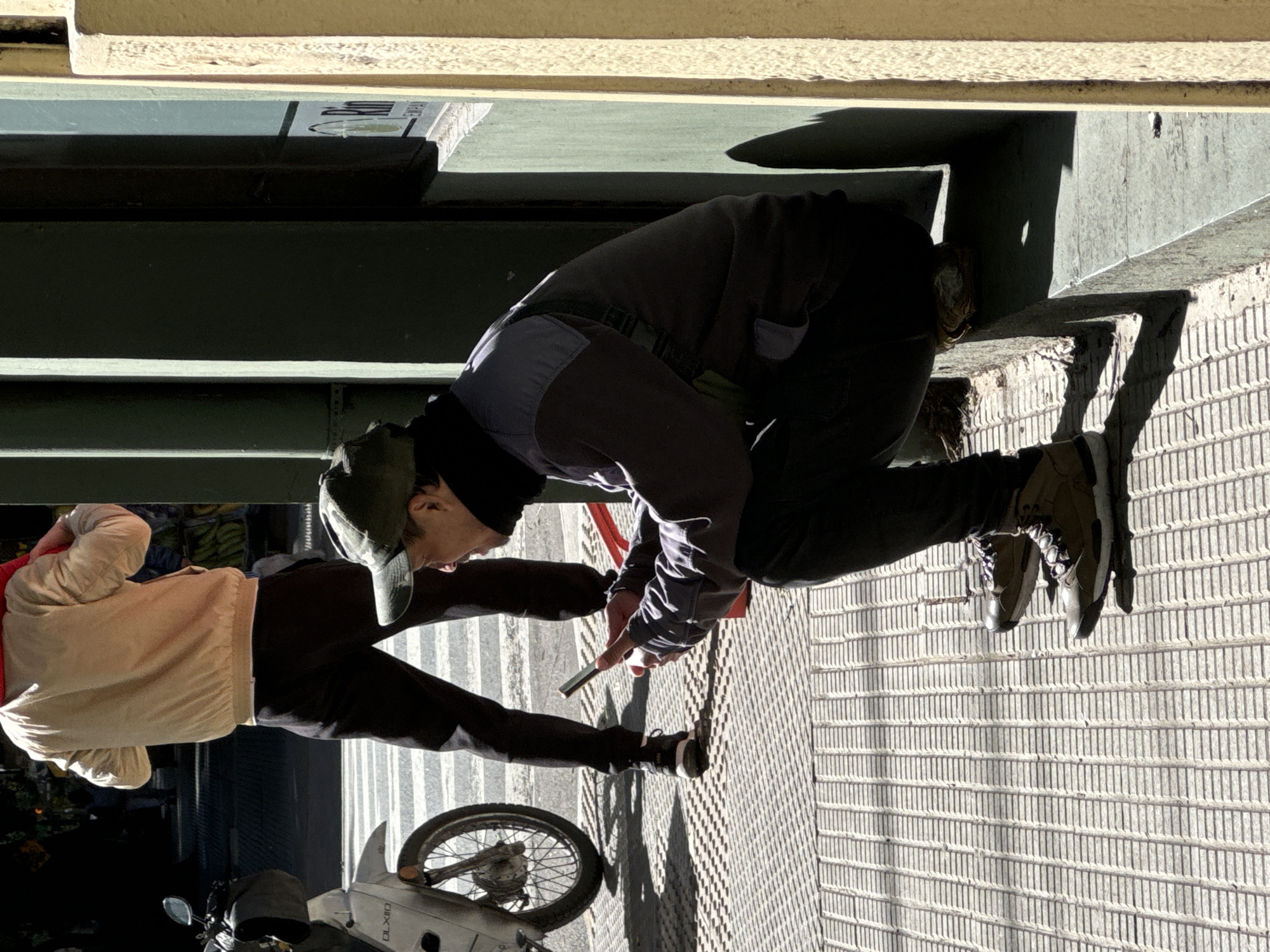 A daytime shot depicts two individuals in an urban setting. One person, wearing a light jacket, dark trousers, and a cap, is standing near a scooter, while the other person kneels and holds a cellphone. The scene appears to be on a sidewalk or pedestrian area next to a building. The background includes a bridge or overpass structure. The location may be in Buenos Aires, Argentina.