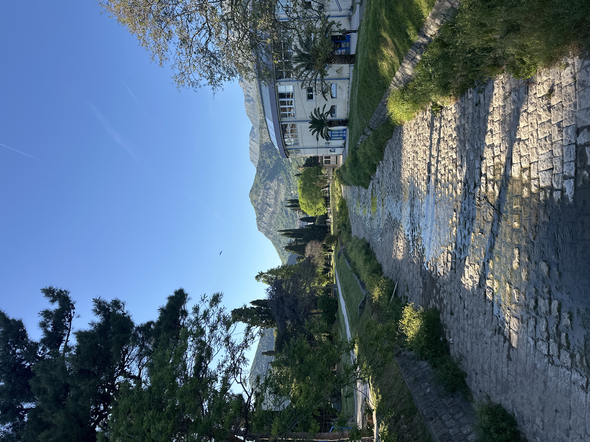 The image captures a low-angle, landscape-oriented view in the city of Budva, Montenegro. A shallow stream or canal with a stone bed runs through the center of the frame, reflecting sunlight. The banks of the waterway are lined with grass and low stone walls. A building with a modern architectural style is visible to the right, partially obscured by trees and featuring a curved section with windows. In the background, a mountain range rises against a clear blue sky. Various trees populate the foreground and mid-ground, casting shadows. A bird is visible in the sky.