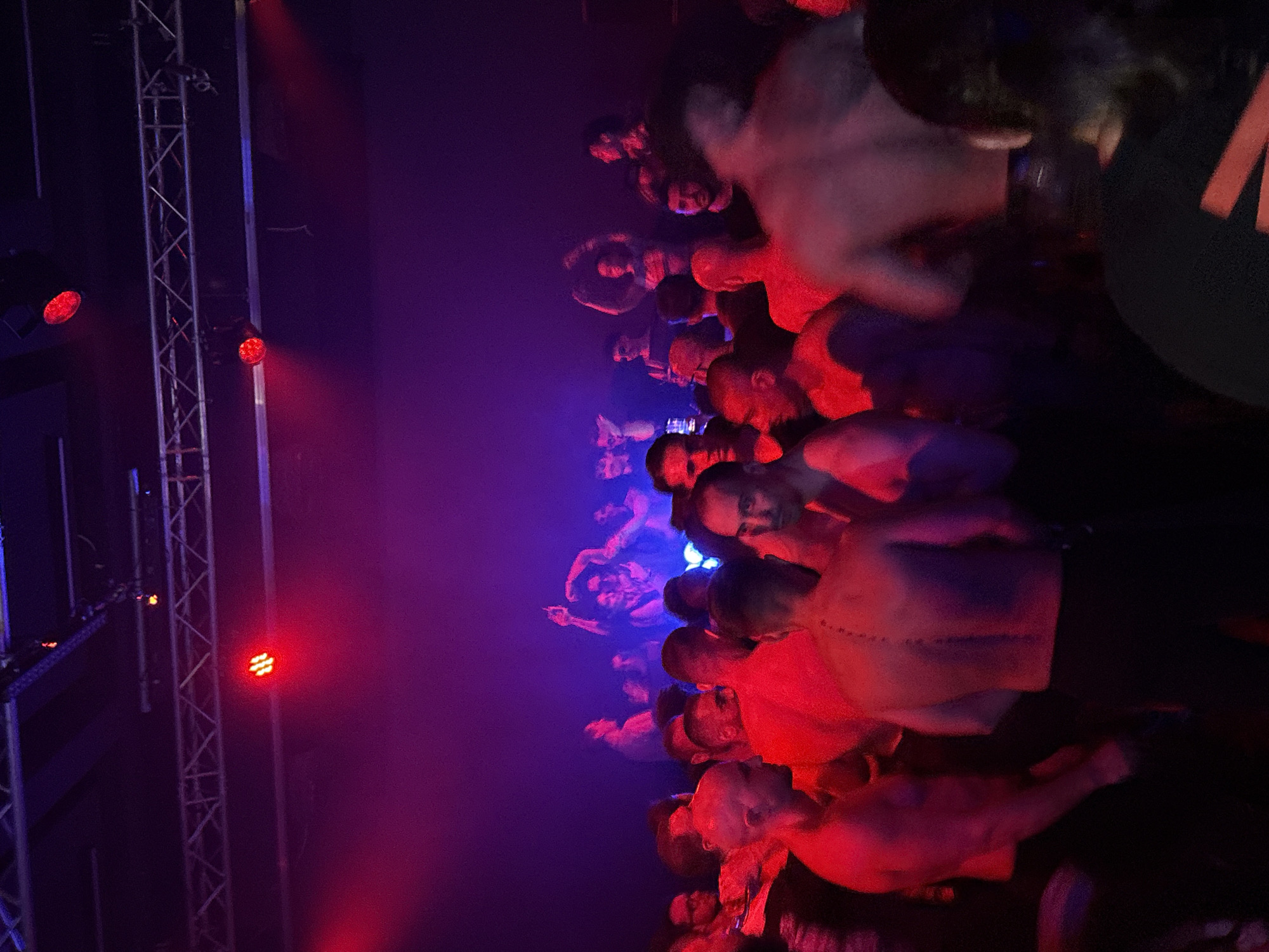 A dimly lit nightclub or performance venue in Sofia, Bulgaria, is filled with a crowd of predominantly male attendees. Red and purple lighting saturates the scene, creating a vibrant and energetic atmosphere. Many individuals are shirtless, with some captured in mid-conversation or dancing. In the background, a stage or elevated area is visible with more people, some appearing to be performers or part of a DJ setup, bathed in blue and white light. The overall impression is one of a lively social gathering or event.