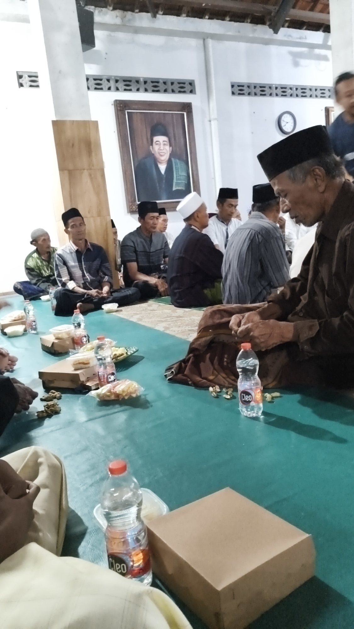 The image depicts an indoor gathering of men, likely a religious or cultural event, taking place in a room with a green carpeted floor in Dalegan, Indonesia. In the background, a framed portrait of a distinguished-looking man in traditional attire hangs on the wall, above a patterned decorative element. Several men are seated on the floor, many wearing peci hats, suggesting Islamic cultural context. Some are engaged in conversation, while others appear to be observing or waiting.
In the foreground, a table-like arrangement on the floor displays several items. These include cardboard food boxes, small packets of snacks, and bottles of water, one clearly labeled "Cleo." Scattered around these items are small, brown nuts or seeds. The lighting suggests an indoor setting, possibly during the day or with artificial illumination. The overall atmosphere appears to be one of quiet assembly and communal gathering.