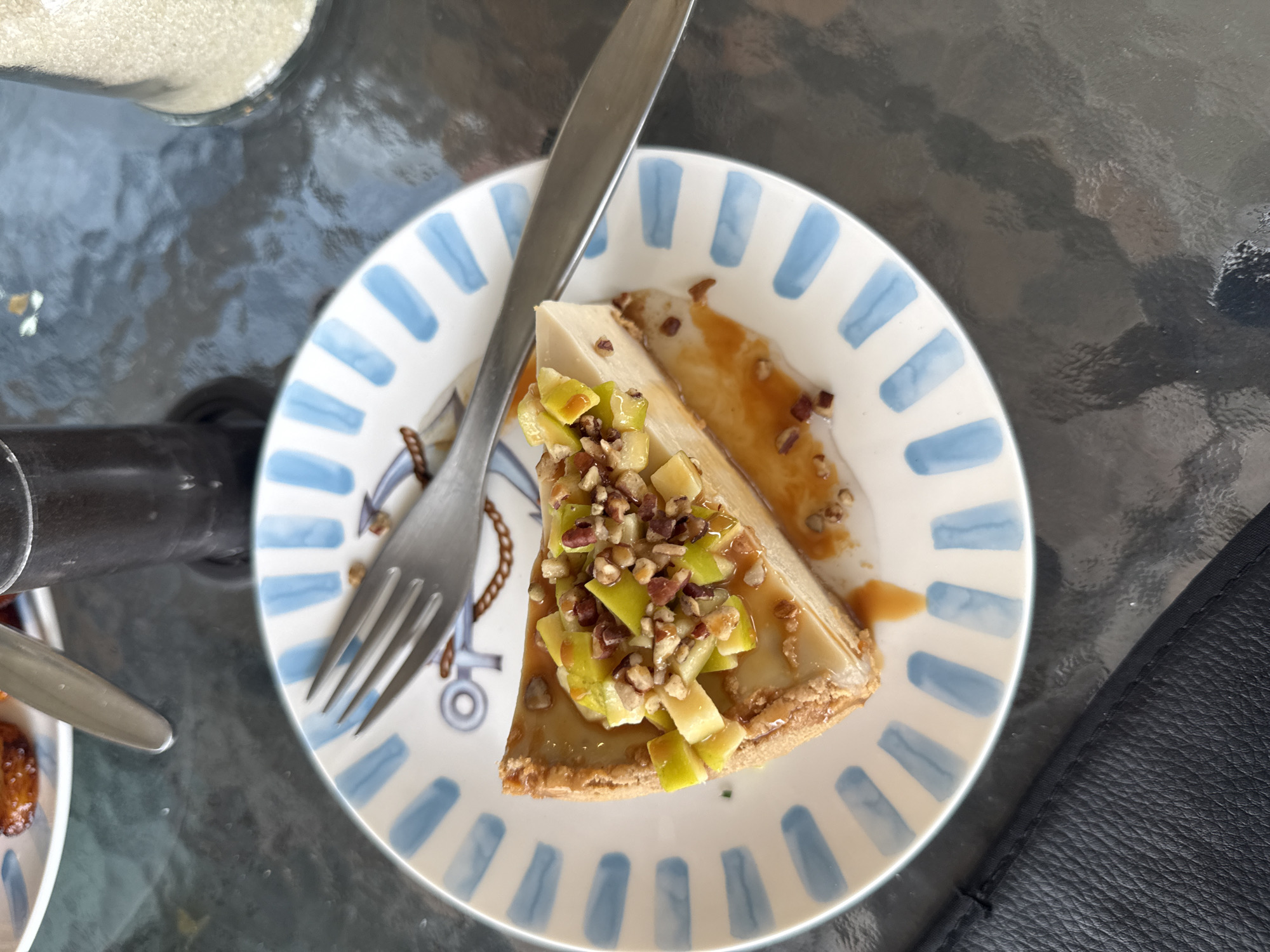 A high-angle shot captures a slice of what appears to be caramel cheesecake, adorned with chopped green apples and nuts, resting on a white plate with blue stripes and nautical anchor motifs. A fork and knife are placed on top of the dessert. The scene is set on a glass-topped table. The location cannot be determined from the image.