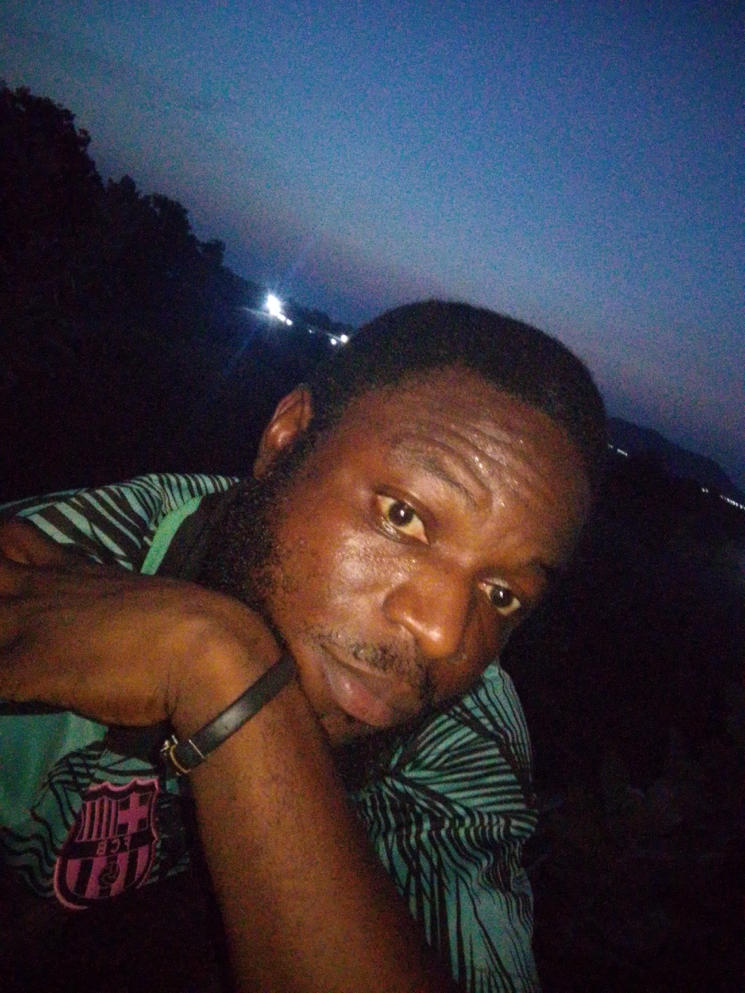 A dark, low-angle close-up shot captures a man with dark skin wearing a blue and black striped shirt. The man is positioned with his arm bent, resting his head on his hand. He is looking directly at the camera. The shirt appears to have a small design or logo on the lower left portion. In the background, distant lights suggest an urban setting, potentially the city of Lokoja, Nigeria. The overall scene is dimly lit.