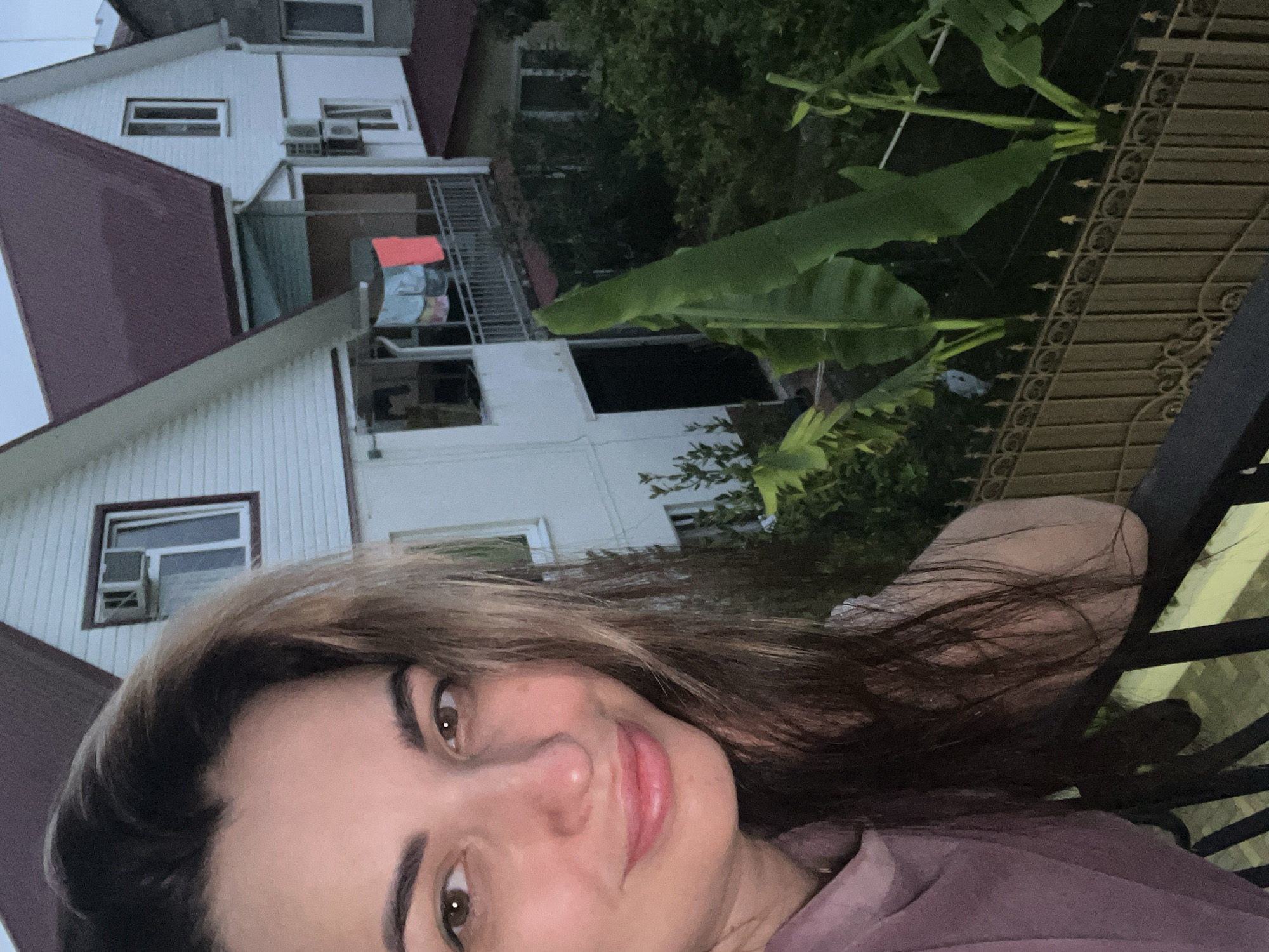 A medium close-up, low-angle selfie shows a young woman with brown hair and brown eyes smiling. She is wearing a purple shirt and leaning on a railing with a fence and a building behind her. The building appears to be a multi-story house with a dark red roof. There are plants in the foreground, including large green leaves, possibly from a banana tree. The lighting suggests it is either early morning or late evening. The location is described as Unknown, Unknown.