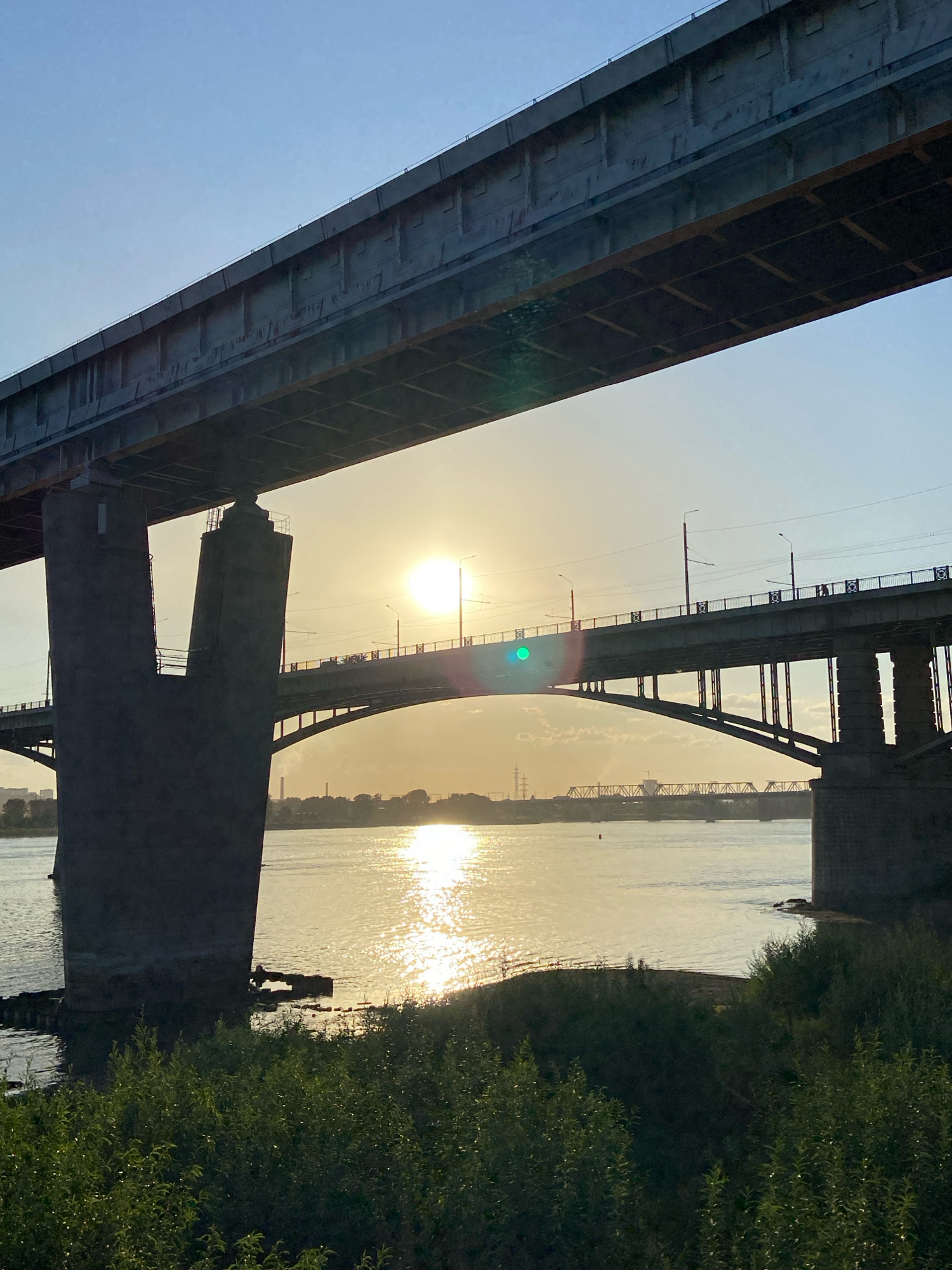 The image depicts a wide river with two bridges spanning it under a bright, hazy sky at sunset. The foreground is dominated by lush green foliage, obscuring the riverbank. The closest bridge is a substantial concrete structure, with one of its massive, dark gray pillars visible on the left. Above this, another, wider bridge stretches horizontally across the frame.
Further in the distance, a second, arched bridge carries traffic, its silhouette and support structures visible against the warm glow of the setting sun, which is positioned slightly to the right of the frame's center. The sun's reflection creates a shimmering path of light across the water. Beyond the second bridge, the faint outlines of city buildings and industrial structures can be seen along the far bank. The atmosphere is serene, with a sense of urban development integrated into a natural riverine landscape.
This scene is likely the Ob River in Novosibirsk, Russia, given the presence of multiple large bridges characteristic of the city's crossings over the river. The specific bridge configuration, particularly the presence of two large bridges in close proximity, suggests a location in the central part of the city, possibly near the Akademgorodok or the city center itself.