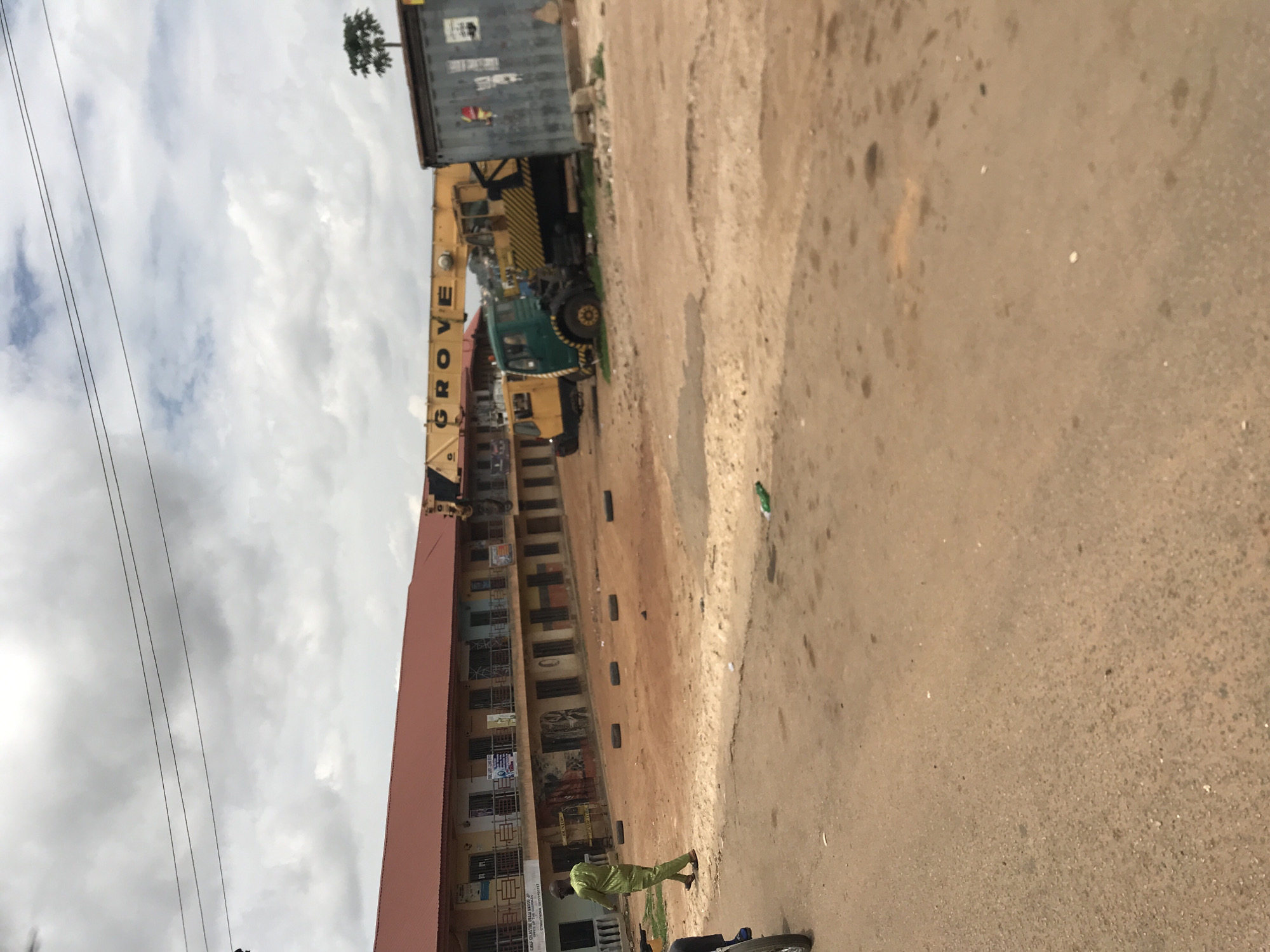 This image depicts a daytime scene in Rafin Par, Nigeria, under an overcast sky with dense grey and white clouds. The foreground consists of a reddish-brown, unpaved road and a large dirt area.
On the left, a man dressed in a light green long-sleeved shirt and matching trousers walks from left to right across the dirt path, his back mostly facing the camera. His feet are visible, wearing sandals. Behind him and further to the left, a section of a vehicle wheel is partially visible.
In the mid-ground, a long, two-story building with a reddish-brown corrugated metal roof extends across much of the frame. The upper floor features a continuous balcony with dark metal railings. The ground floor has multiple storefronts or entrances, some with signage. Visible text on one sign includes "CHIEF CELESTINE URASU", "0706...", and "0805...". Several black tires are laid flat in a line on the dirt in front of this building.
To the right of the building, a large yellow mobile crane is prominently parked. Its horizontal boom clearly displays the brand name "GROVE" in large black letters. A green truck is positioned directly in front of the crane, partially obscuring its base. Another yellow vehicle is visible behind the green truck.
Further to the right, a grey metal container or storage unit stands, bearing some faint markings and stickers. Above and behind this container, a single, slender tree with foliage at its crown is silhouetted against the sky. Power lines traverse the upper portion of the sky, extending horizontally across the frame. A discarded green plastic bottle is visible on the dirt ground in the lower mid-right.