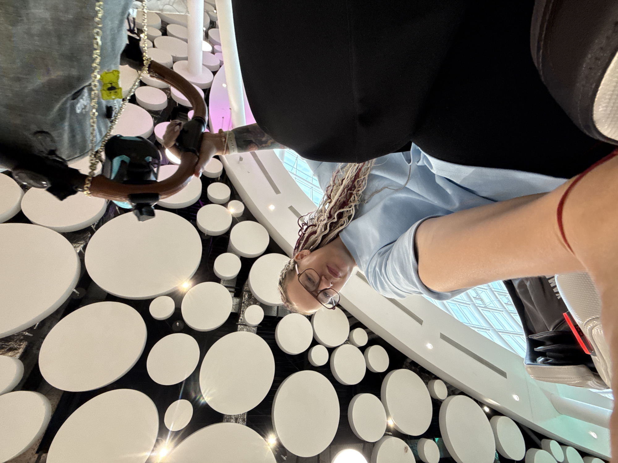 The image shows a low-angle, wide-shot perspective looking up at a woman with blonde and reddish-brown braided hair wearing glasses and a light blue t-shirt. She is seated in a black chair, and a portion of her left arm is visible, along with what appears to be a red string bracelet. Her right arm is holding a device with a wooden handle and a black camera attached to it.
The background features a wall decorated with numerous large, circular white panels of varying sizes, creating a mosaic-like effect. Some of these panels have a subtle sheen, reflecting the lighting. Below the woman, a large section of black fabric is visible, likely part of her clothing or the chair she is in. To the left of the woman, a light-colored section of what might be a massage chair is visible, with a red accent light. In the upper right portion of the image, a person's gray clothing and a black glove are visible, suggesting another person is present, possibly operating the camera or interacting with the woman.
The environment appears to be indoors, possibly a public space or a business. The ceiling above is a dark color, and the white circles are affixed to it. The overall impression is of a modern, stylized interior. No specific Moscow landmarks or indicators are present in the image.