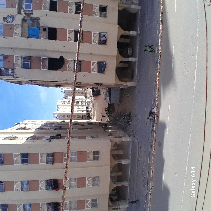 The image captures a devastated urban street scene in Khan Yunis, Palestinian Territory, under a clear blue sky. Two multi-story residential buildings, constructed from light-colored concrete with patterned window surrounds, flank a wide thoroughfare. Both buildings display extensive damage, with numerous broken or missing windows, some covered by makeshift materials like a blue tarp or dark fabric. Visible holes and impact marks scar their facades, notably a large, irregular opening on an upper floor of the building on the left. The ground level between the buildings is filled with debris, rubble, and what appears to be discarded household items, indicating significant destruction. Further down this unpaved area, several small, indistinct figures are visible amidst temporary structures, possibly tents or shacks.
On the right side of the image, a paved asphalt road with distinct white lane markings stretches into the distance. Two individuals are walking on this road, away from the viewer. One person is prominently wearing a bright yellow-green vest over dark clothing, while the other is dressed in dark attire. The ground floor of the building on the right features a series of arched openings, under which damaged vehicles or debris are partially visible. A thick, multi-colored rope or cable runs diagonally across the foreground, slightly obscuring the view. The bright daylight and clear sky suggest the photo was taken during the day, possibly mid-morning or afternoon. The overall atmosphere is one of post-conflict devastation and hardship. The text "Galaxy A14" is visible in the bottom right corner of the image.
