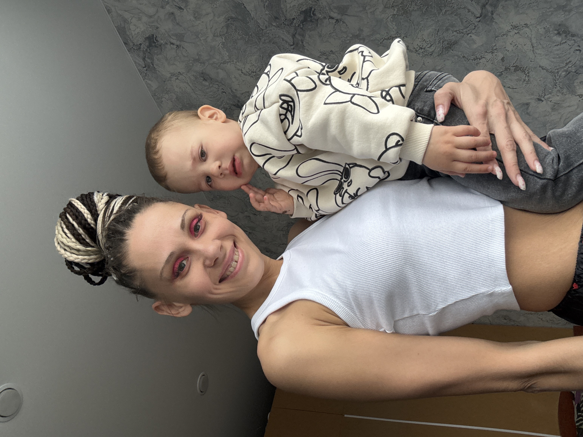 A woman with a braided bun hairstyle is smiling and holding a toddler. The toddler is wearing a cream-colored sweatshirt with black graphics and grey jeans. The woman is wearing a white tank top and pink eye makeup. The background consists of a wall with a grey marble-like pattern and a grey wall. The location cannot be determined as Rusanovo, Russia based on the given information.