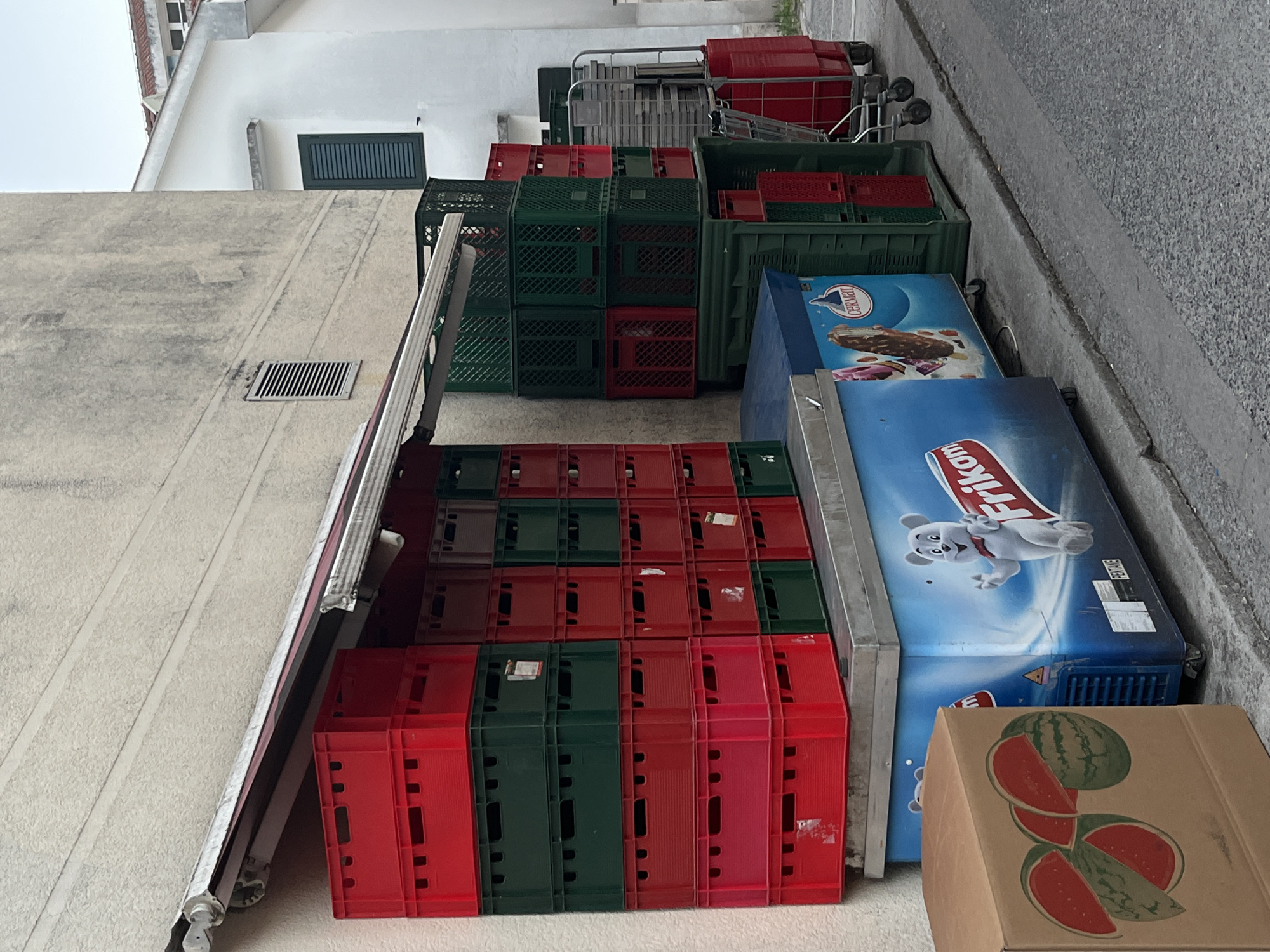 The image captures a street scene in Budva, Montenegro, featuring an outdoor display of various items, likely for sale. In the foreground, a brown cardboard box with images of sliced watermelons is positioned to the left. Adjacent to it, a blue commercial freezer displays advertisements for Frikam ice cream and features an animated polar bear graphic. Stacked high against a textured, light-colored building wall are numerous plastic crates, predominantly in red and green, with some red crates also appearing to the right. Behind these stacks, a white building with a dark green shuttered window is visible. Further to the right, a wheeled metal cart with red and gray plastic crates is parked. The ground consists of paved asphalt and concrete. The overall environment suggests an outdoor market or a retail establishment with overflow merchandise.