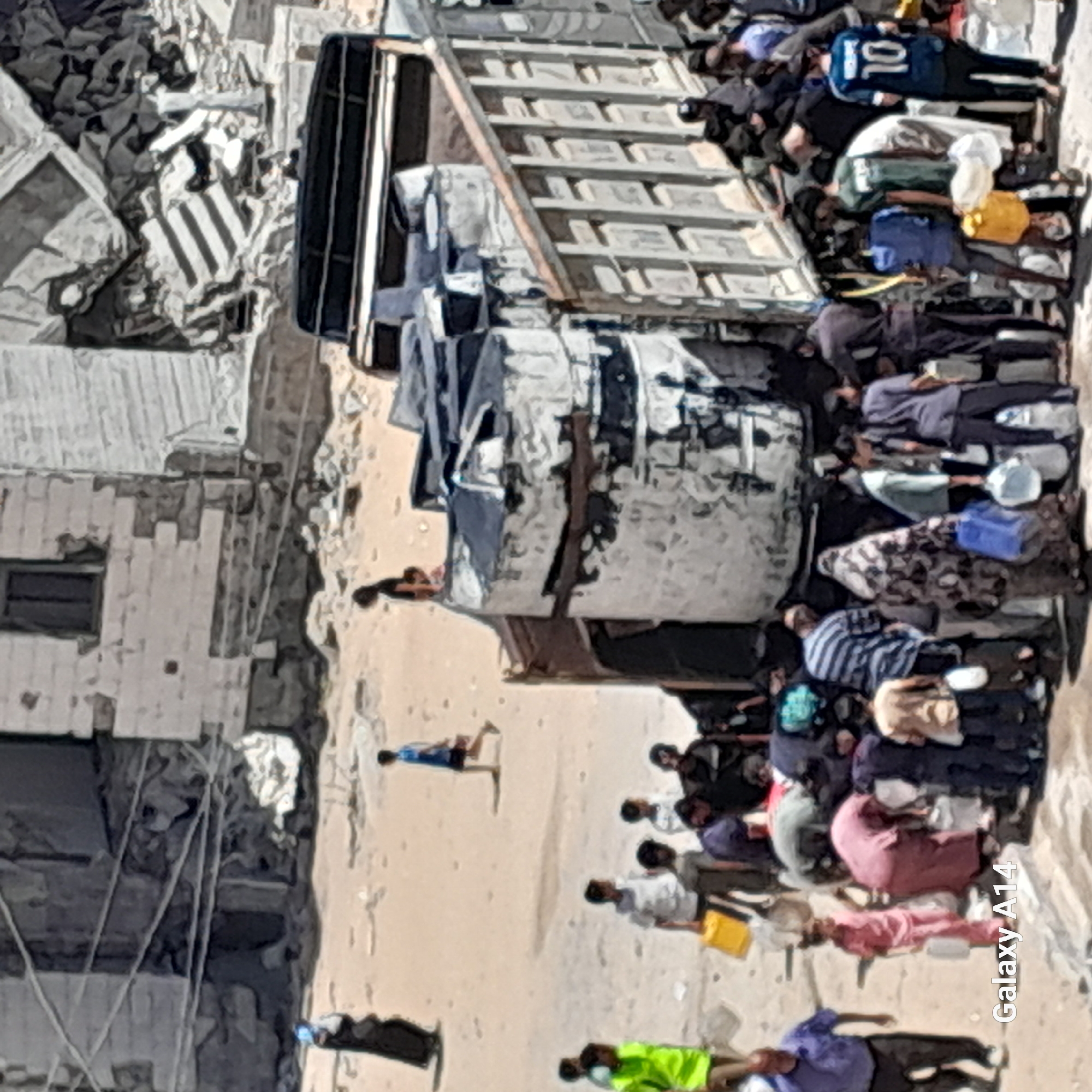 In the city of Deir el-Balah, Palestinian Territories, a group of people are gathered around a truck filled with water containers. The scene appears to be a relief effort, with individuals collecting water. The environment shows signs of damage, with rubble and destroyed buildings in the background. The people, including men, women, and children, are dressed in casual clothing, with some women wearing headscarves. They are seen carrying empty containers and interacting with each other as they wait for their turn to receive water. The truck itself is a large vehicle with multiple large, dark water tanks mounted on its back. The ground is sandy, and there are debris scattered around, suggesting a recent event of destruction. The overall impression is one of a community struggling with scarcity and seeking essential resources amidst a difficult environment.