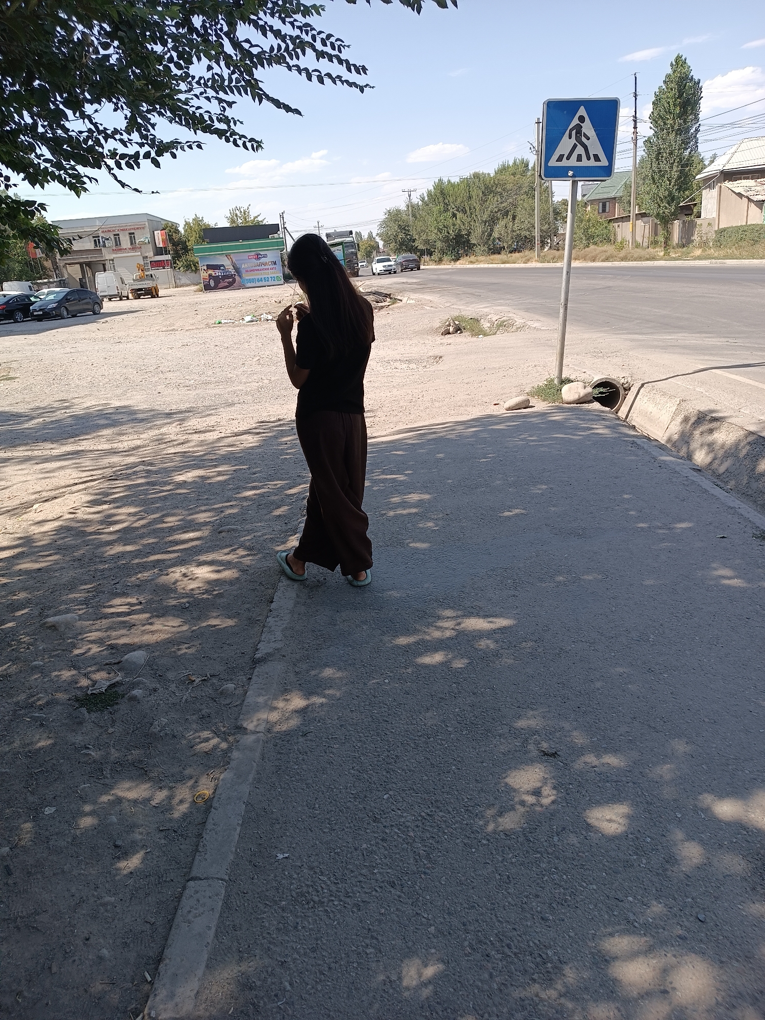The image shows a young woman standing on a sidewalk next to a road in Bishkek City, Kyrgyzstan. The woman is wearing a dark-colored t-shirt and loose brown pants, with light-colored sandals. She is facing away from the camera and appears to be looking at her phone or something in her hands. The sidewalk is made of asphalt and has a concrete curb. The road is paved and has traffic lanes. There are trees and buildings visible in the background, as well as a pedestrian crossing sign. The sky is clear and blue. The lighting suggests it is daytime.