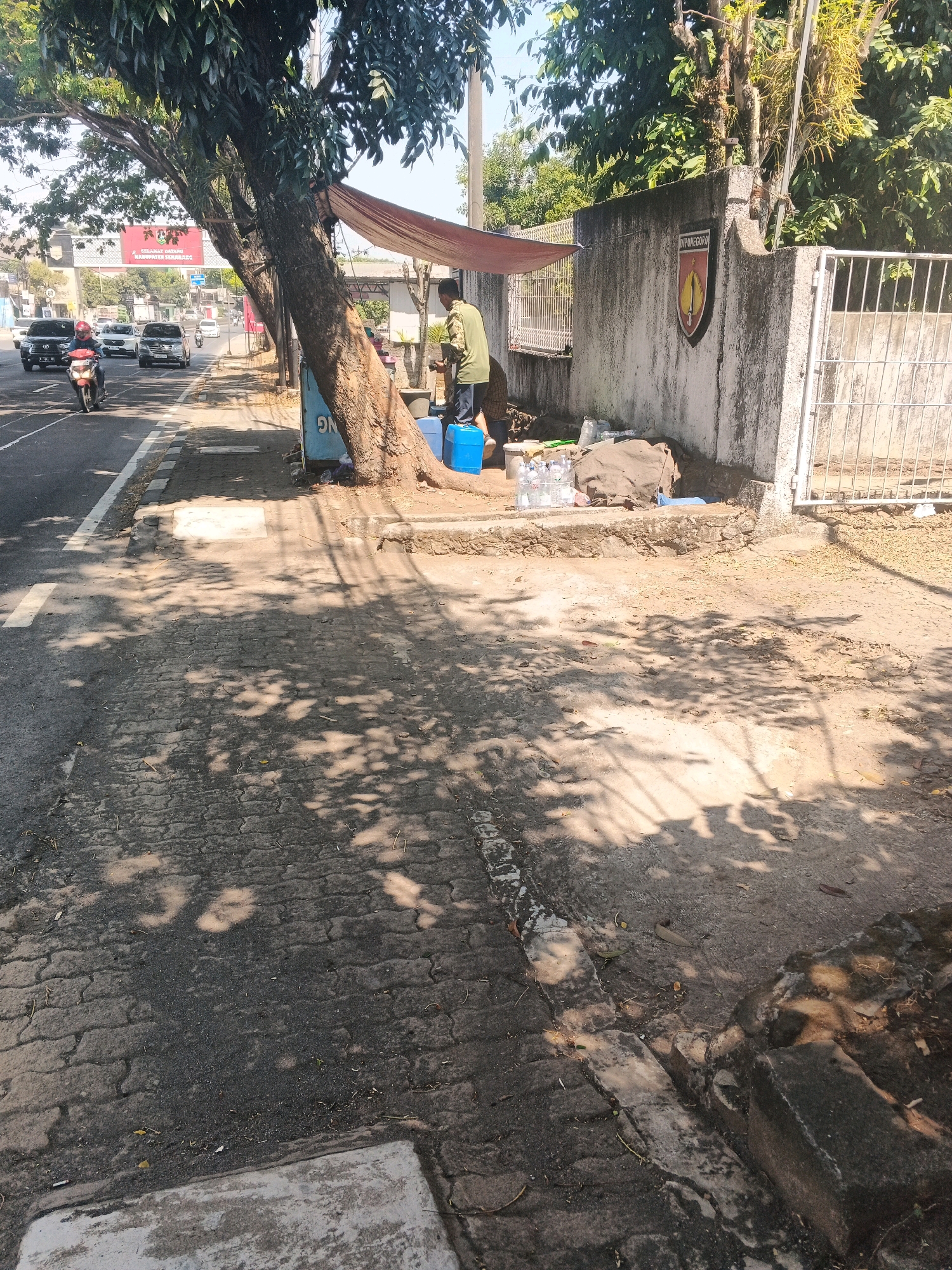 The image depicts a street scene in Semarang, Indonesia, featuring a sidewalk adjacent to a road with moderate traffic. A large tree shades a portion of the sidewalk. On the sidewalk, a person wearing a green shirt and dark pants stands near a blue plastic container and several bottles of water. A tarp is stretched over some of the items. To the right, a concrete wall with a crest bearing the inscription "DIPONEGORO" is visible, along with a metal fence. The ground on the sidewalk is paved with interlocking bricks, and the road is asphalt. The overall environment suggests a sunny day, with tree shadows dappling the ground.