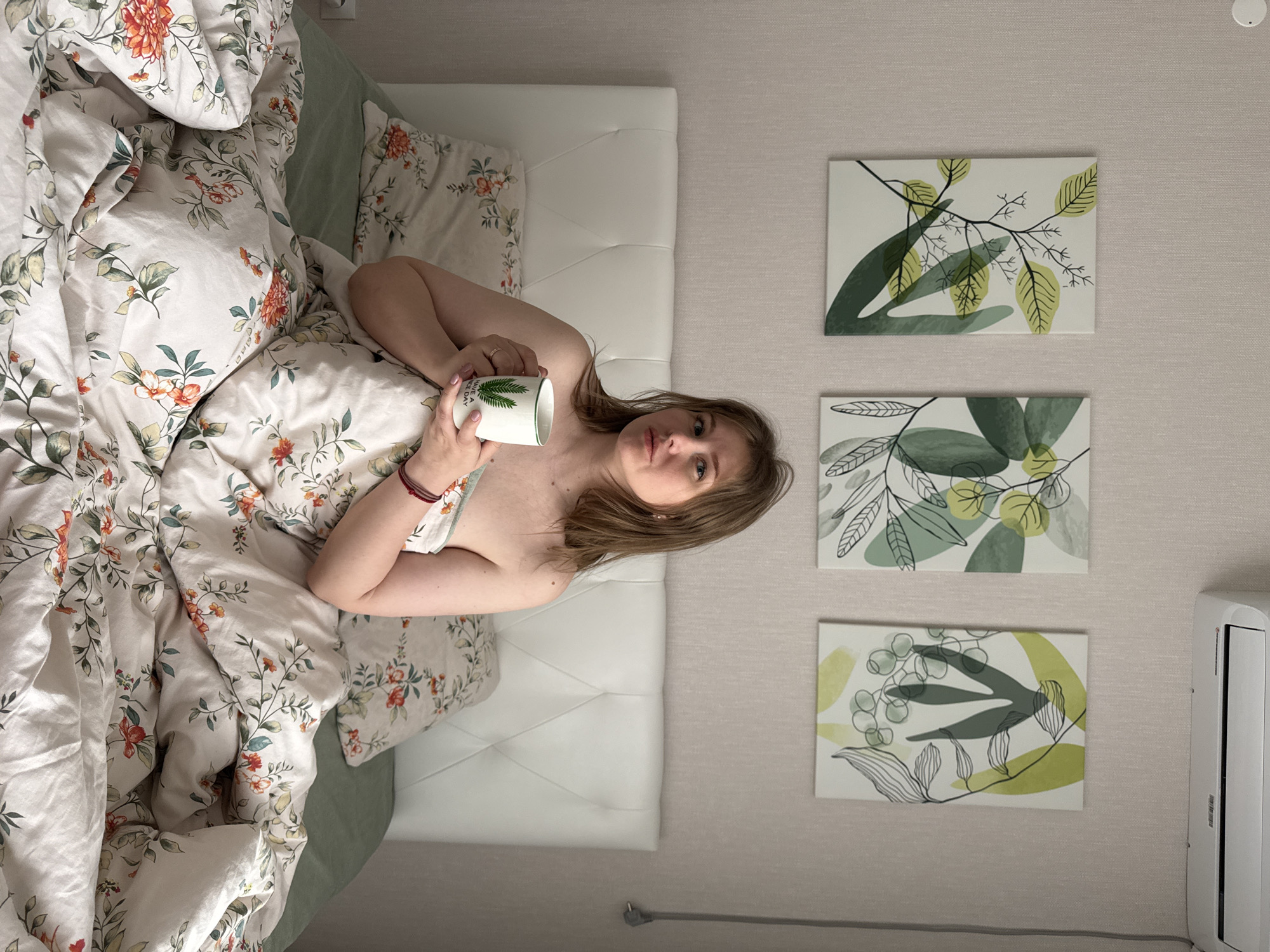 An overhead shot shows a young woman relaxing in bed, facing the camera with a neutral expression. She lies on her side, partially covered by a white duvet adorned with an intricate floral pattern of orange blossoms and green leaves. She holds a white mug with a green leaf design and the partially visible text "HAVE A NICE DAY" in her right hand. A red bracelet is on her left wrist, and a ring is on her right hand. Her light brown hair is spread around her head, and her shoulders are bare. The bed has a white padded headboard. To the right of the bed, on a light-colored, textured wall, three rectangular artworks featuring abstract green and yellow botanical designs are hung vertically. Below the lowest artwork, a white air conditioning unit is mounted. The scene is set indoors, likely a bedroom in Perm, Russia.