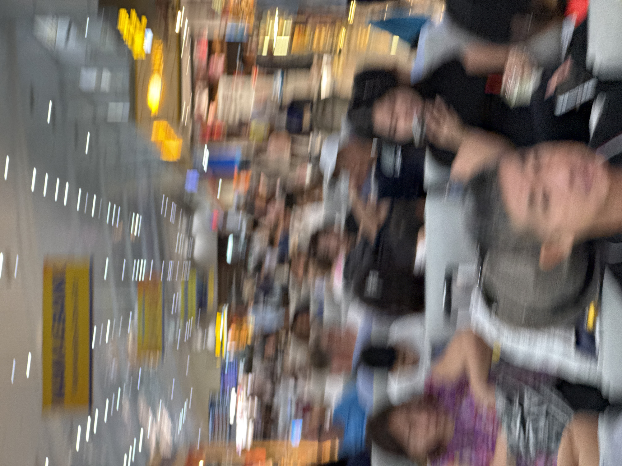 The image shows a blurred interior scene with several people sitting in rows of chairs, likely in an airport terminal or waiting area. Yellow signs with text are visible in the background, one of which appears to be an directional sign. The overall impression is of a busy public space with people waiting. The location is identified as Bac Cam Ranh Ward, Vietnam.